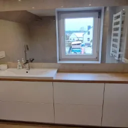 Nowoczesna łazienka z białymi szafkami pod umywalką z drewnianym blatem, duże lustro i grzejnik drabinkowy na szarej ścianie, widok z okna na zabudowę jednorodzinną.