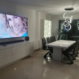 Nowoczesna jadalnia z białym stołem, krzesłami z czarnej ekoskóry na metalowych nogach, białą szafką RTV pod telewizorem i wiszącą lampą z okręgów, w tle witryna i bieżnia.