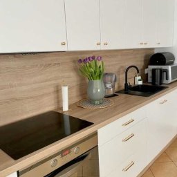 GutArt Radosław Gut - Nowoczesna biała kuchnia z drewnianym blatem, minimalistyczne szafki z podłużnymi uchwytami, płyta indukcyjna i piekarnik ze stali nierdzewnej, dekoracyjny wazon z fioletowymi tulipanami.