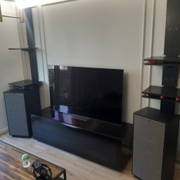 GutArt Radosław Gut - Czarna szafka RTV z połyskiem, telewizor, głośniki i półki wiszące na ścianie z dekoracyjnymi listwami, widok na drewniany stolik kawowy z akcesoriami.