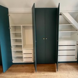 GutArt Radosław Gut - Zestaw szaf wnękowych z ciemnozielonymi frontami i złotymi gałkami, z otwartymi drzwiami ukazującymi białe wnętrze z półkami i szufladami, dopasowany do skosu dachu.