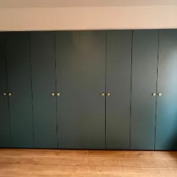 Długa, ciemnozielona szafa z ośmioma drzwiami i okrągłymi, złotymi uchwytami, dopasowana do skosu dachu, na drewnianej podłodze w jasnym wnętrzu.