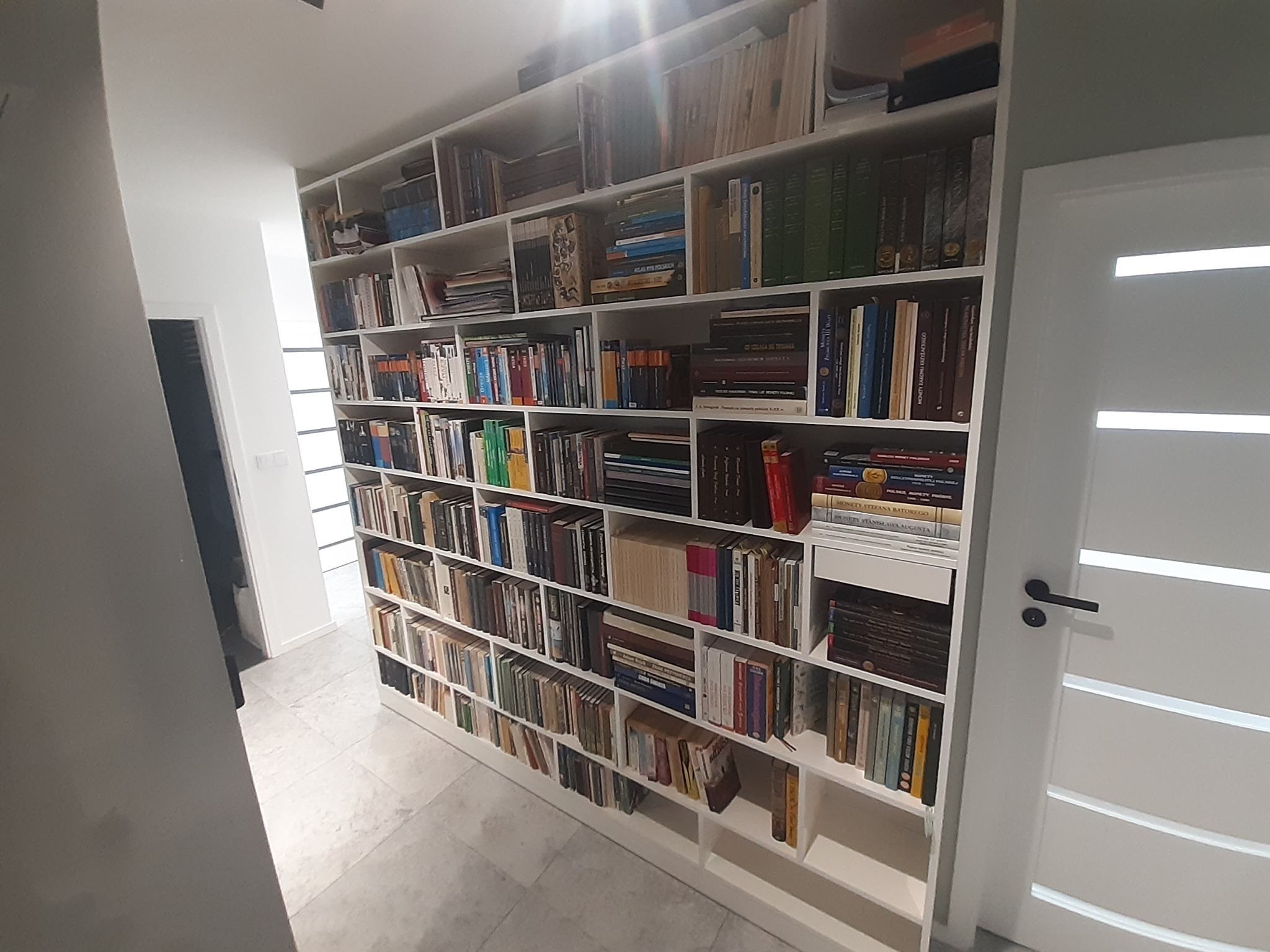 Biała, wysoka zabudowa regałowa na książki, wypełniająca całą ścianę, z widocznym oświetleniem górnym i marmurową podłogą w tle