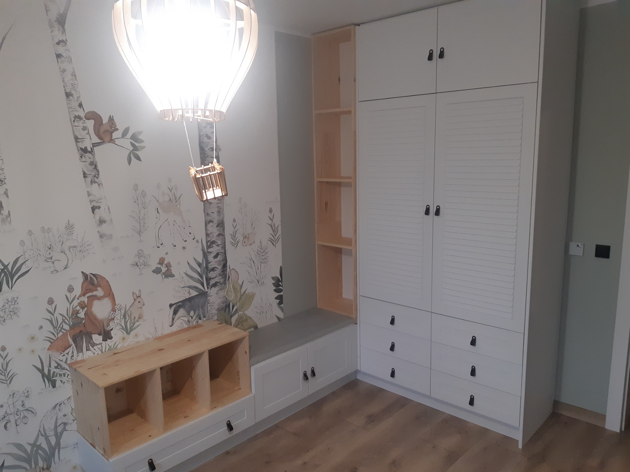 Biała szafa z lamelowymi drzwiami i szufladami w pokoju dziecięcym z tapetą w leśne zwierzęta, uzupełniona o drewniany regał i siedzisko z szarą poduszką.