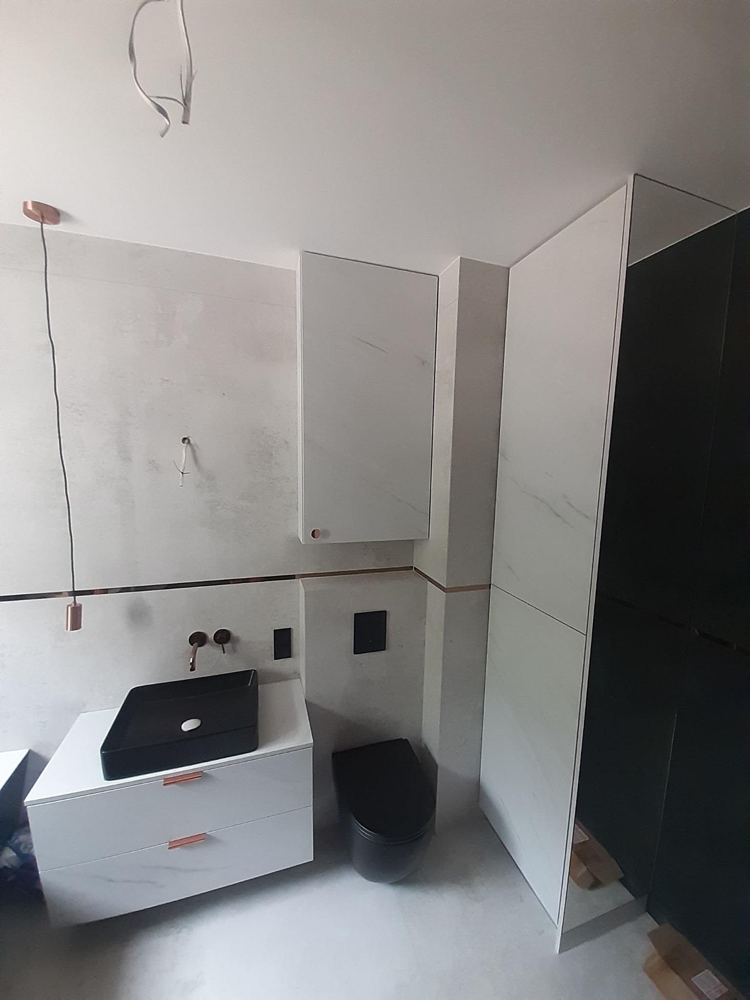 Nowoczesna łazienka z białą szafką pod umywalkę z czarną umywalką nablatową, wiszącą szafką i czarną toaletą, wykończona miedzianymi akcentami i oświetleniem, w trakcie montażu.