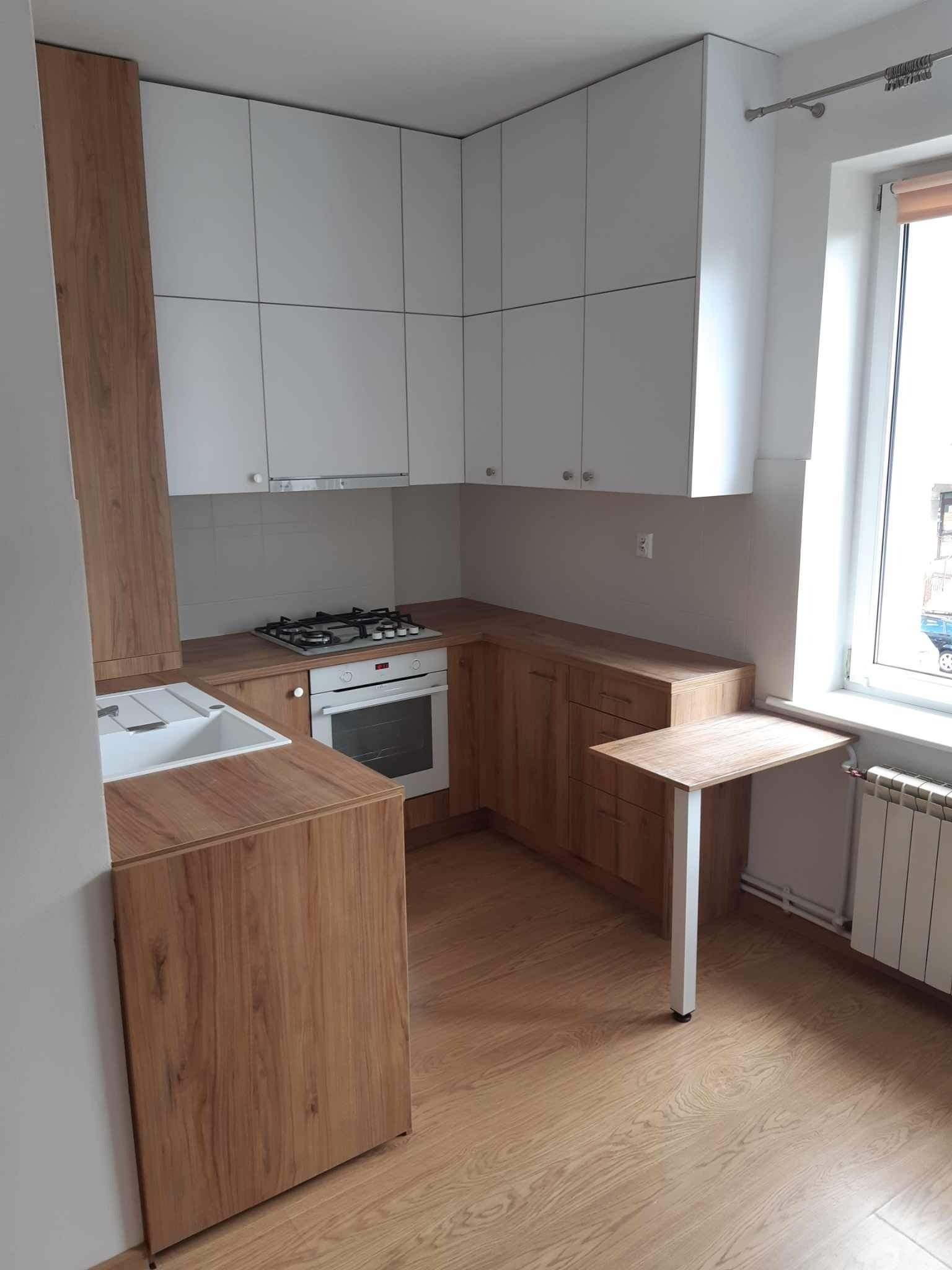 Jasna kuchnia z białymi szafkami górnymi i drewnopodobnymi szafkami dolnymi, z blatem i małym rozkładanym stolikiem przy oknie.