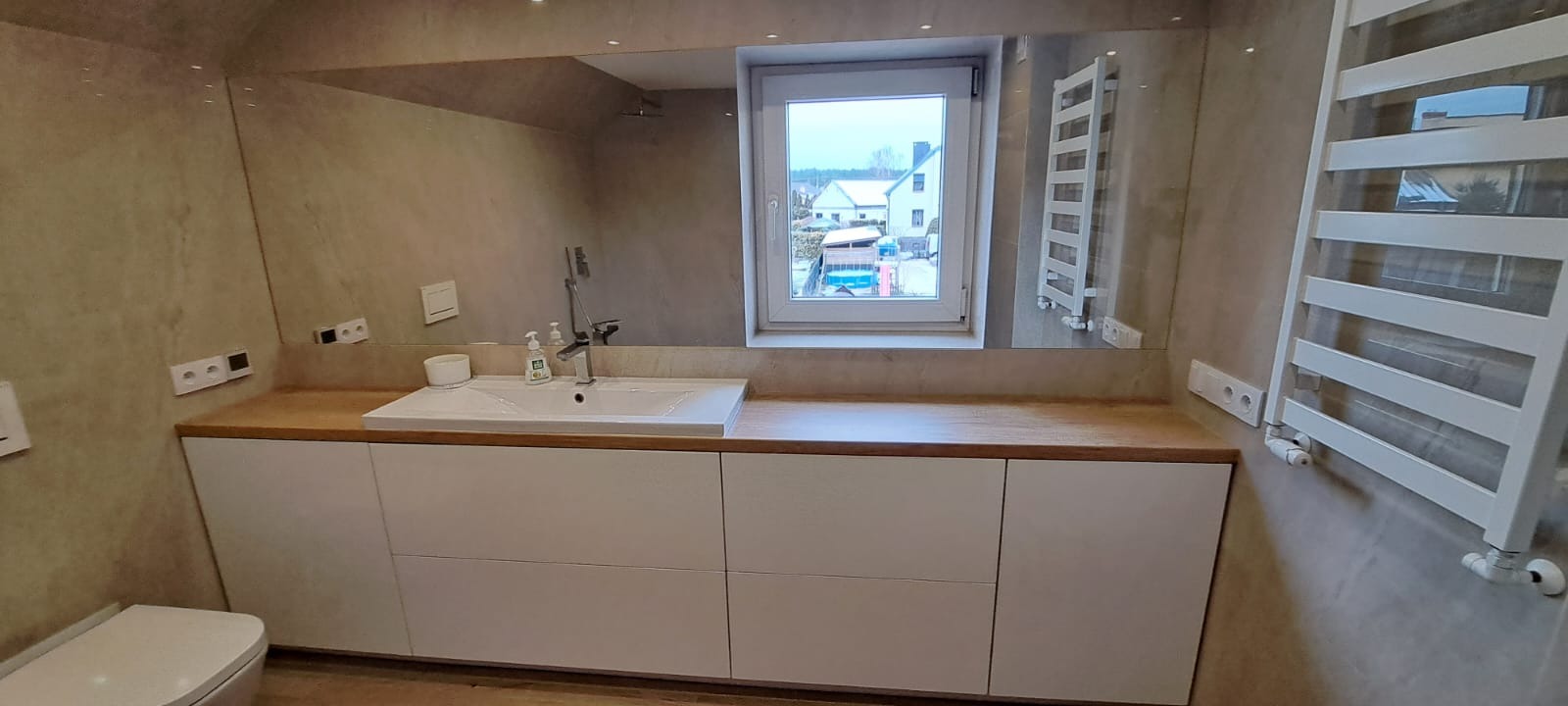 Nowoczesna łazienka z białymi szafkami pod umywalką z drewnianym blatem, duże lustro i grzejnik drabinkowy na szarej ścianie, widok z okna na zabudowę jednorodzinną.