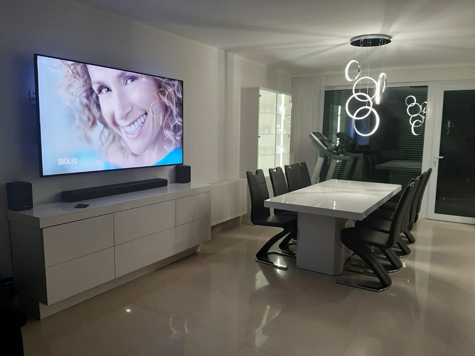 Nowoczesna jadalnia z białym stołem, krzesłami z czarnej ekoskóry na metalowych nogach, białą szafką RTV pod telewizorem i wiszącą lampą z okręgów, w tle witryna i bieżnia.