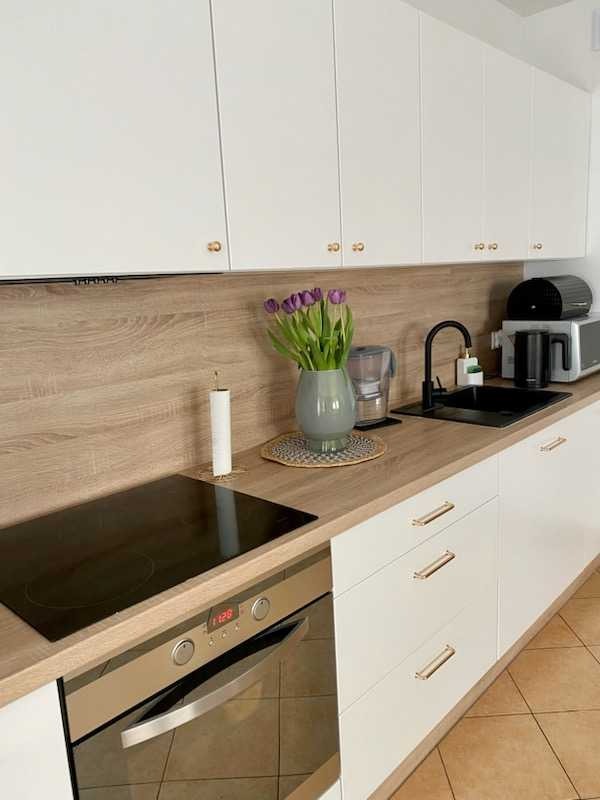 Nowoczesna biała kuchnia z drewnianym blatem, minimalistyczne szafki z podłużnymi uchwytami, płyta indukcyjna i piekarnik ze stali nierdzewnej, dekoracyjny wazon z fioletowymi tulipanami.