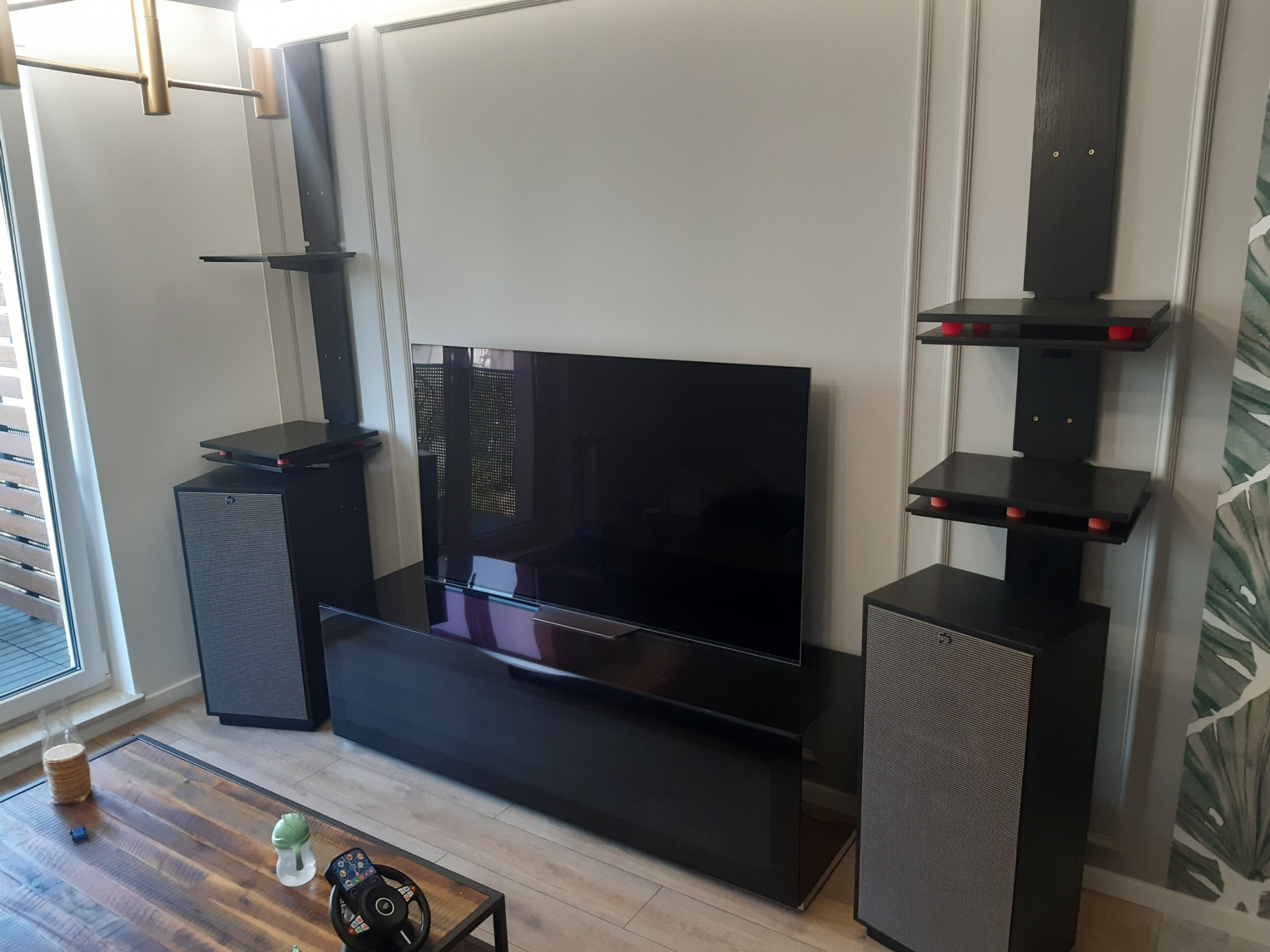 Czarna szafka RTV z połyskiem, telewizor, głośniki i półki wiszące na ścianie z dekoracyjnymi listwami, widok na drewniany stolik kawowy z akcesoriami.