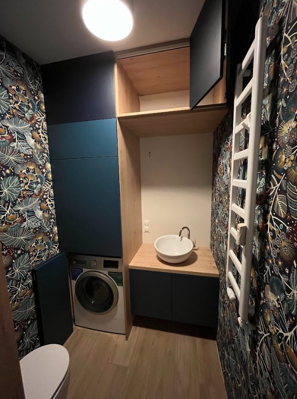 Niebiesko-drewniana zabudowa łazienkowa z pralką, umywalką nablatową i grzejnikiem drabinkowym na tle wzorzystej tapety.