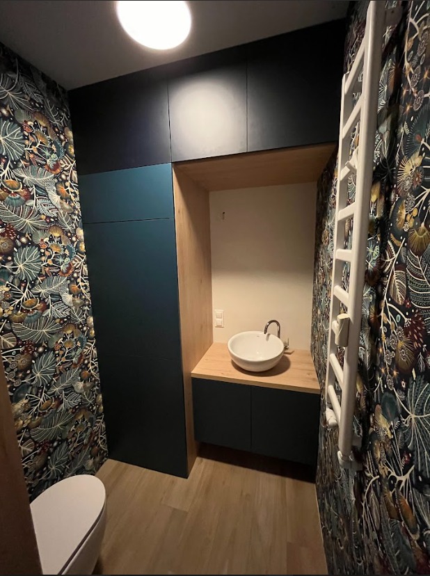 Mała łazienka z zabudową meblową w odcieniach granatu i drewna, wzorzysta tapeta na ścianach, okrągła umywalka nablatowa i grzejnik drabinkowy.