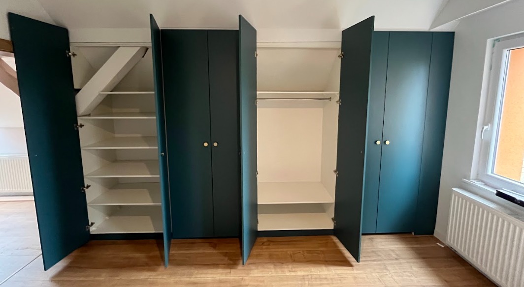 Zabudowa wnęki z szafami w kolorze ciemnej zieleni, z otwartymi drzwiami ukazującymi białe wnętrze z półkami i drążkiem, dopasowana do skosu dachu.