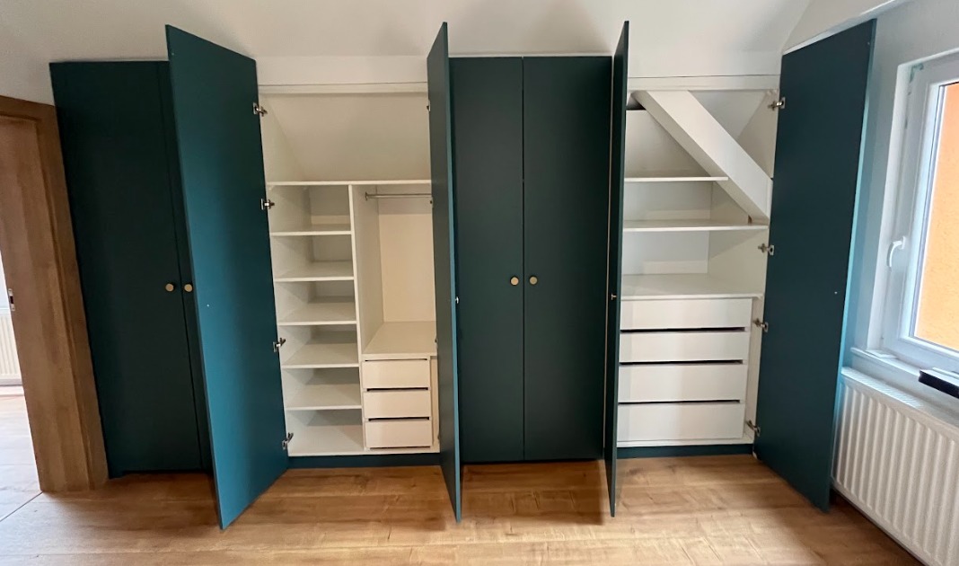 Zestaw szaf wnękowych z ciemnozielonymi frontami i złotymi gałkami, z otwartymi drzwiami ukazującymi białe wnętrze z półkami i szufladami, dopasowany do skosu dachu.