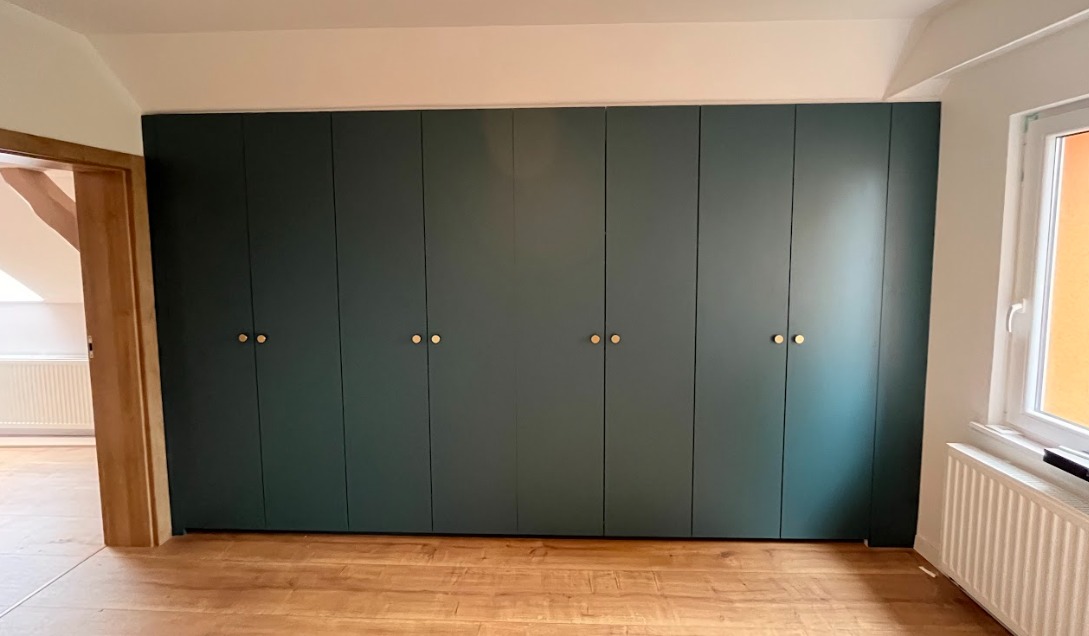 Długa, ciemnozielona szafa z ośmioma drzwiami i okrągłymi, złotymi uchwytami, dopasowana do skosu dachu, na drewnianej podłodze w jasnym wnętrzu.