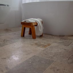 Wit-bud - Nowoczesna łazienka z wolnostojącą białą wanną, drewnianym stołkiem z ręcznikiem i podłogą z płytek imitujących naturalny kamień w odcieniach szarości i beżu.