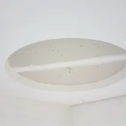 Owalny otwór w suficie z płyt kartonowo-gipsowych, widoczne mocowania i przewody elektryczne, sufit w trakcie wykańczania.