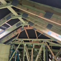 Drewniana konstrukcja dachu z oknem połaciowym, wzmocniona metalowymi łącznikami, zabezpieczona folią. Widoczne poziomice i laserowy niwelator.