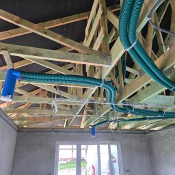 Wit-bud - Drewniana konstrukcja dachu z zamontowanymi, zielonymi rurami wentylacyjnymi i instalacją elektryczną w surowym wnętrzu budynku. Widoczne okno i częściowo otynkowane ściany.