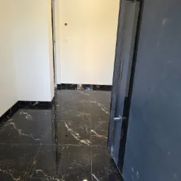 Połyskująca, czarna marmurowa podłoga w korytarzu z białymi ścianami. Widoczna część otwartych, szarych drzwi z metalową klamką. Nowoczesne wykończenie.