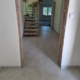 Wit-bud - Wnętrze w trakcie prac wykończeniowych: szare płytki podłogowe, fragment schodów zabezpieczonych folią, ściany z odsłoniętą cegłą przy przejściu.