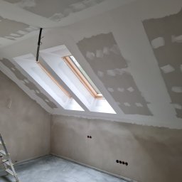 Wit-bud - Pokój na poddaszu w trakcie prac wykończeniowych: ściany i sufit po szpachlowaniu, widoczne okna dachowe i instalacja elektryczna. Fragment drabiny w lewym dolnym rogu.
