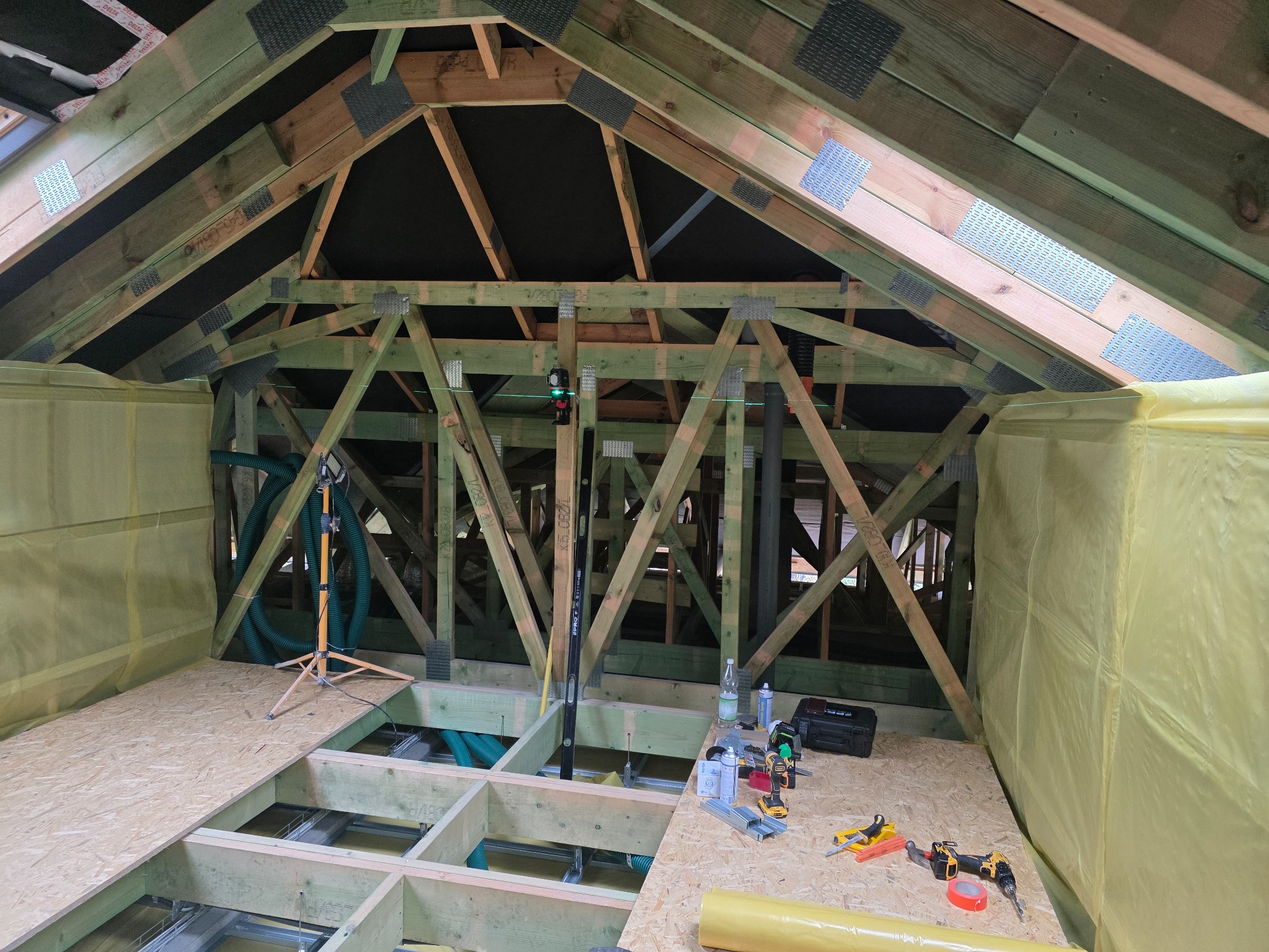 Poddasze w trakcie adaptacji: drewniana konstrukcja dachu ze wzmocnieniami, poziomica laserowa, narzędzia i materiały budowlane. Widoczne etapy prac wykończeniowych.