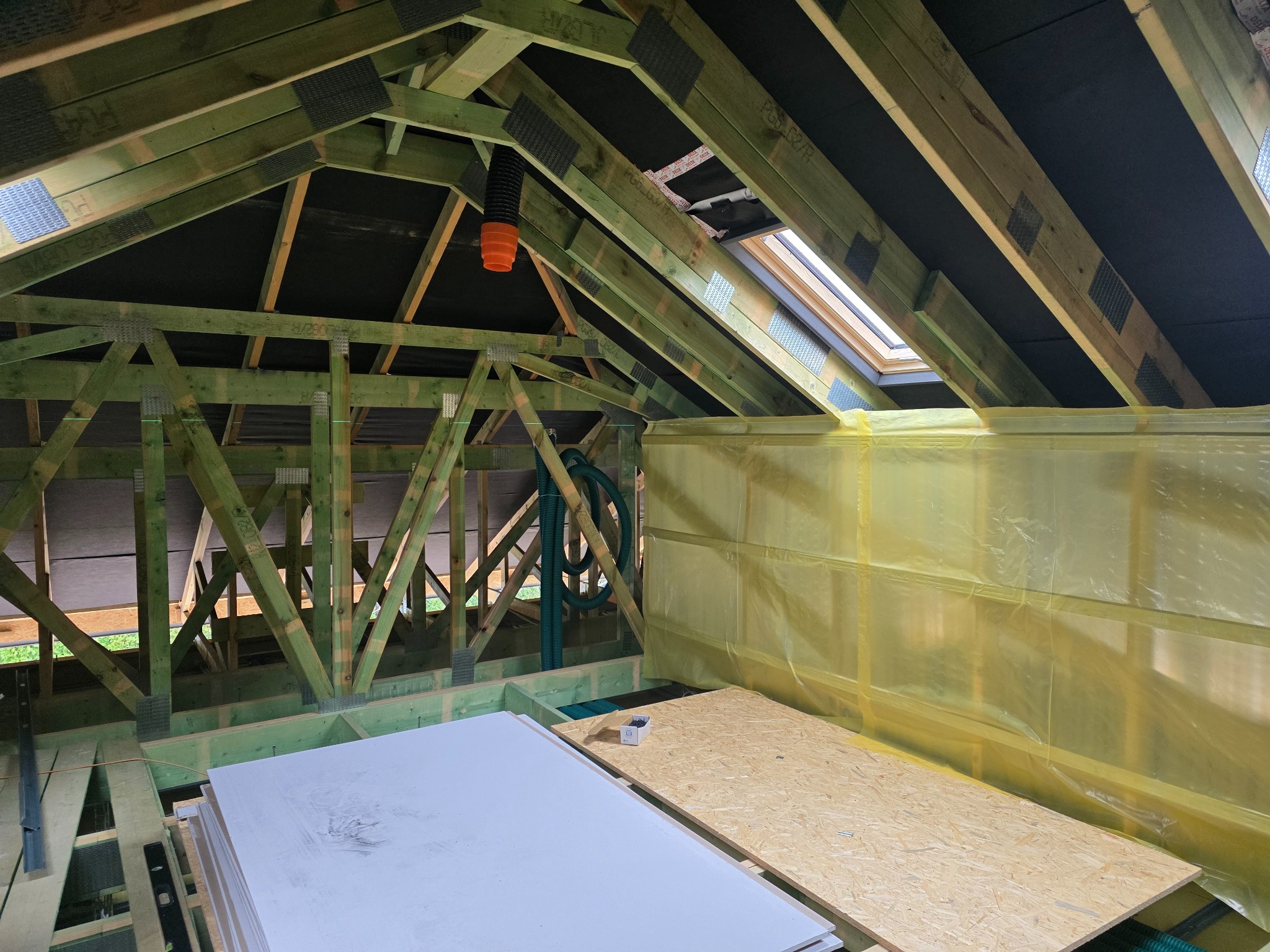 Konstrukcja dachu z oknem połaciowym, widoczne płyty kartonowo-gipsowe i elementy drewniane, zabezpieczone folią. Prace wykończeniowe na poddaszu w trakcie realizacji.