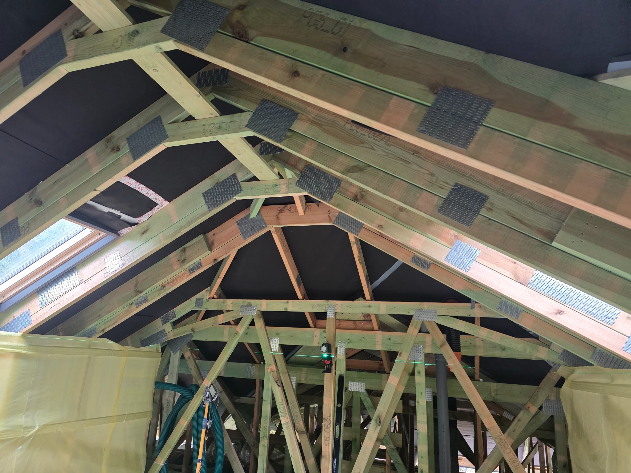 Drewniana konstrukcja dachu z oknem połaciowym, wzmocniona metalowymi łącznikami, zabezpieczona folią. Widoczne poziomice i laserowy niwelator.
