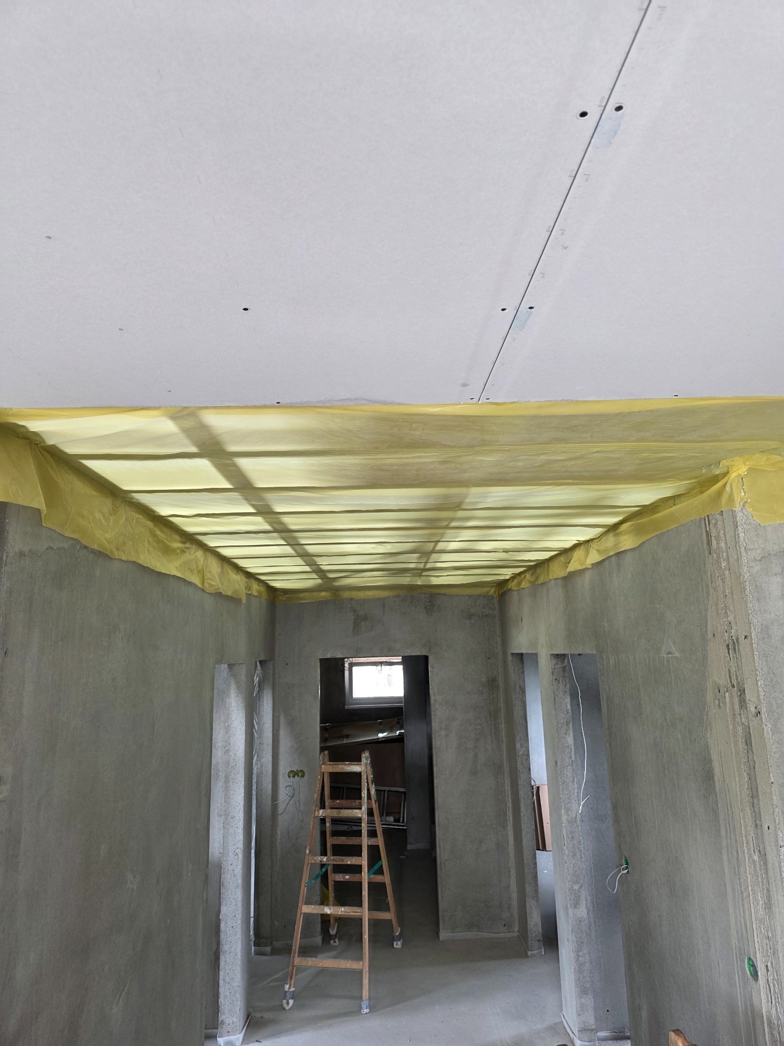 Sufit podwieszany z płyt karton-gips, zabezpieczony folią ochronną, widoczna drabina w przejściu. Surowe ściany w tle. Prace wykończeniowe w trakcie.