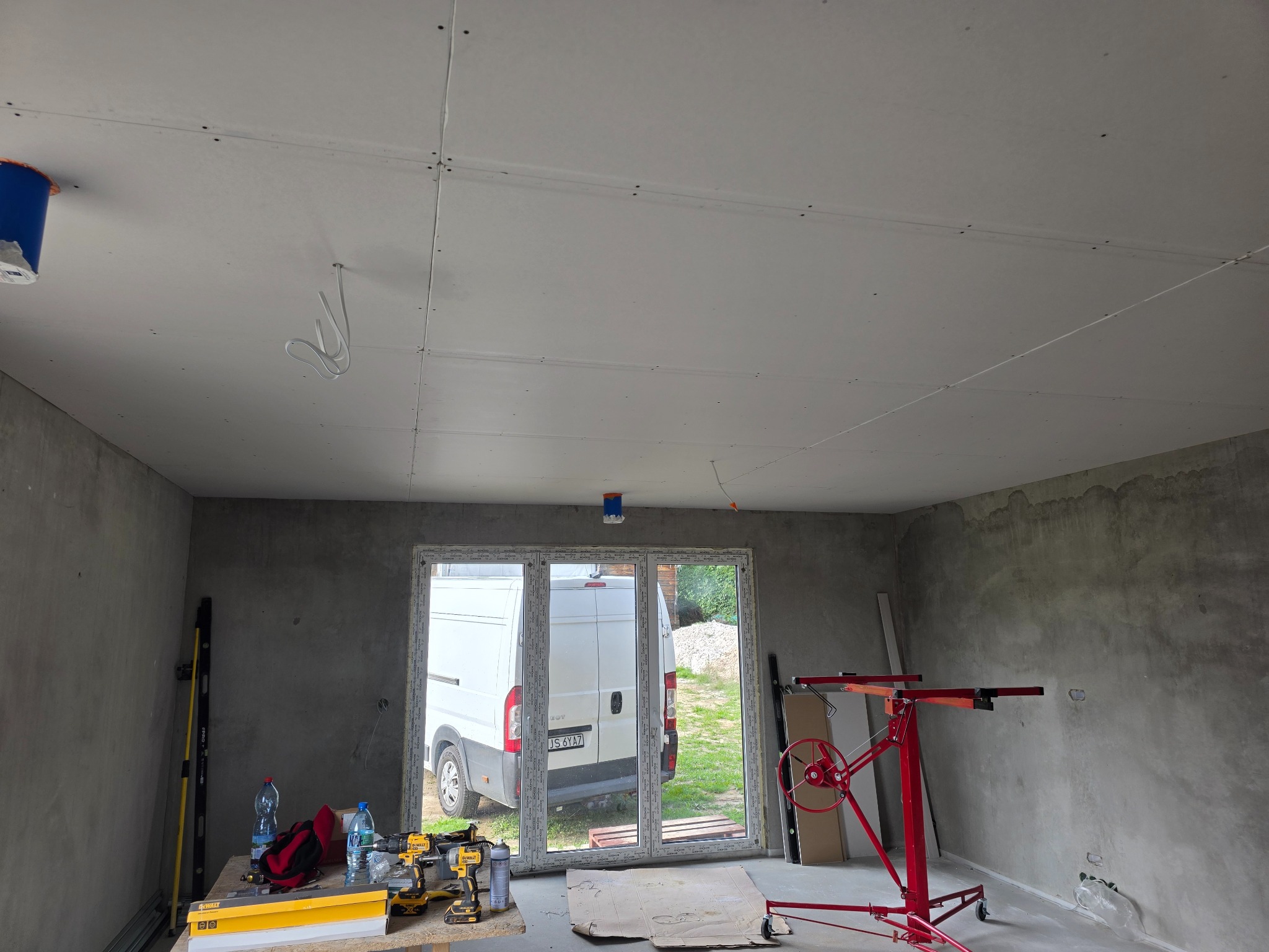 Montaż płyt karton-gips na suficie w surowym wnętrzu. Widoczny podnośnik do płyt, narzędzia i częściowo widoczny samochód dostawczy za oknem.