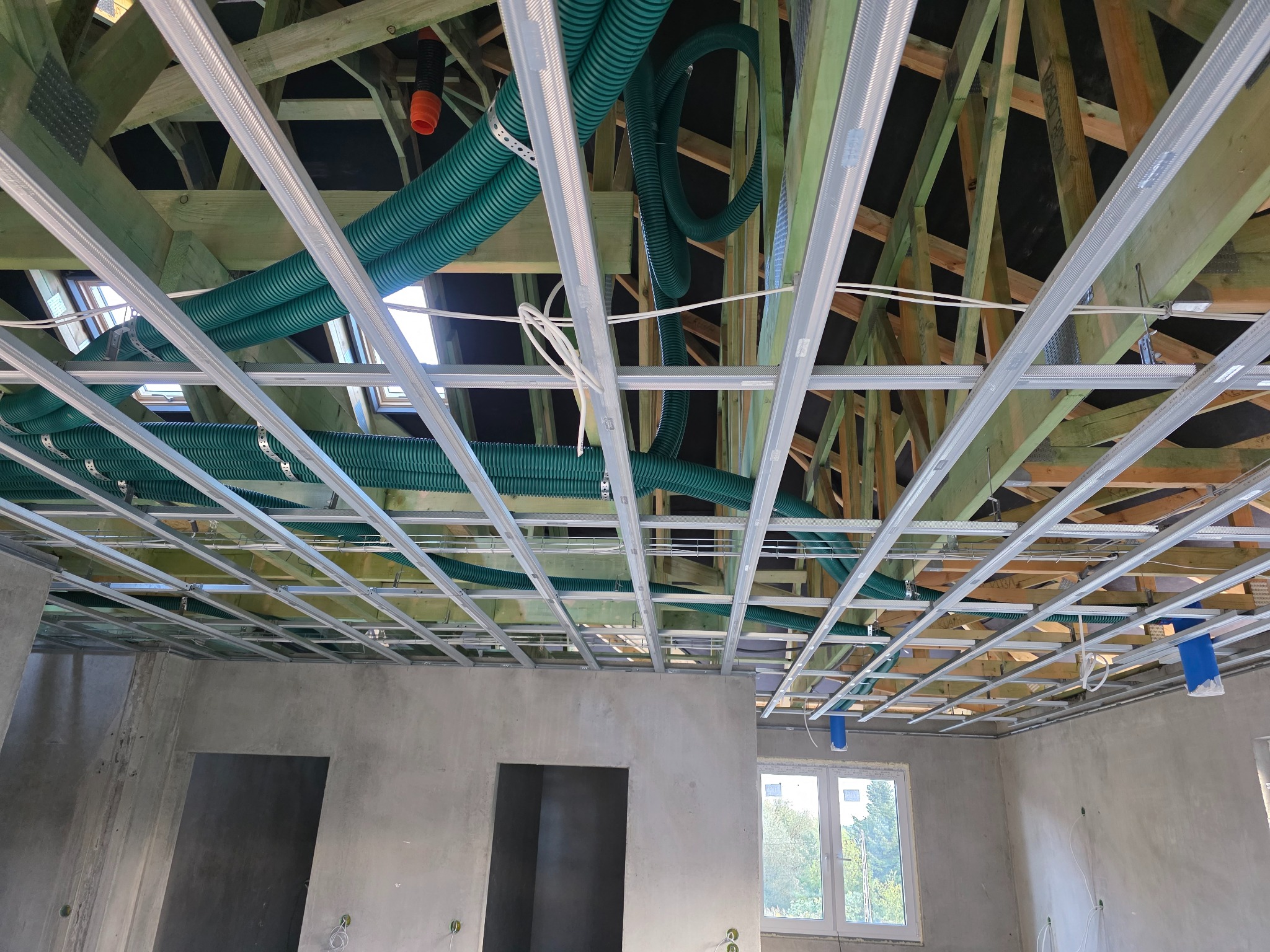 Montaż konstrukcji sufitu podwieszanego z widoczną instalacją wentylacyjną w surowym wnętrzu. Widoczne profile i zielone rury wentylacyjne.