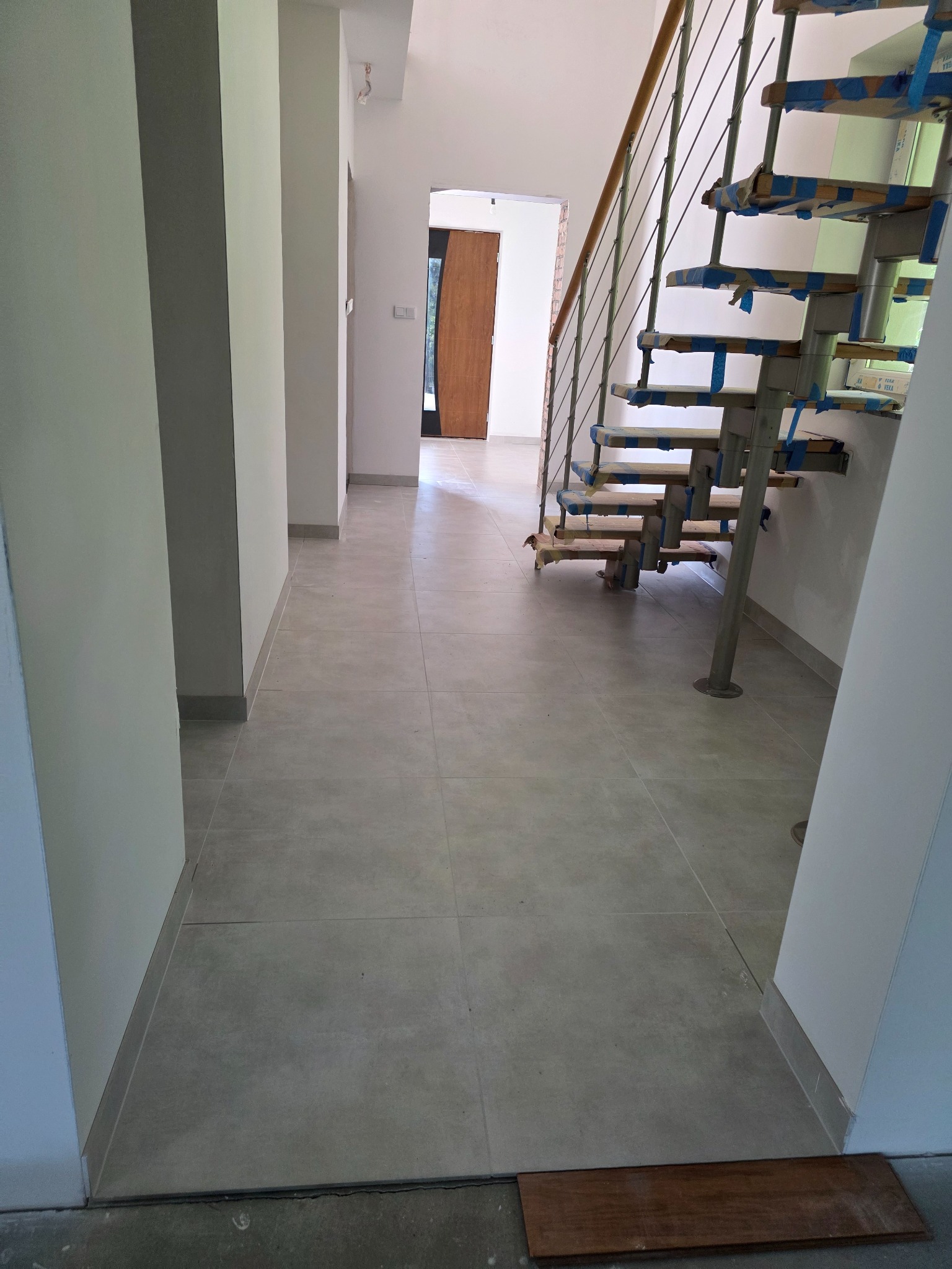 Jasne płytki podłogowe w korytarzu z minimalistycznymi schodami z metalowymi elementami, zabezpieczonymi niebieską taśmą. Widok na drzwi wejściowe w tle.