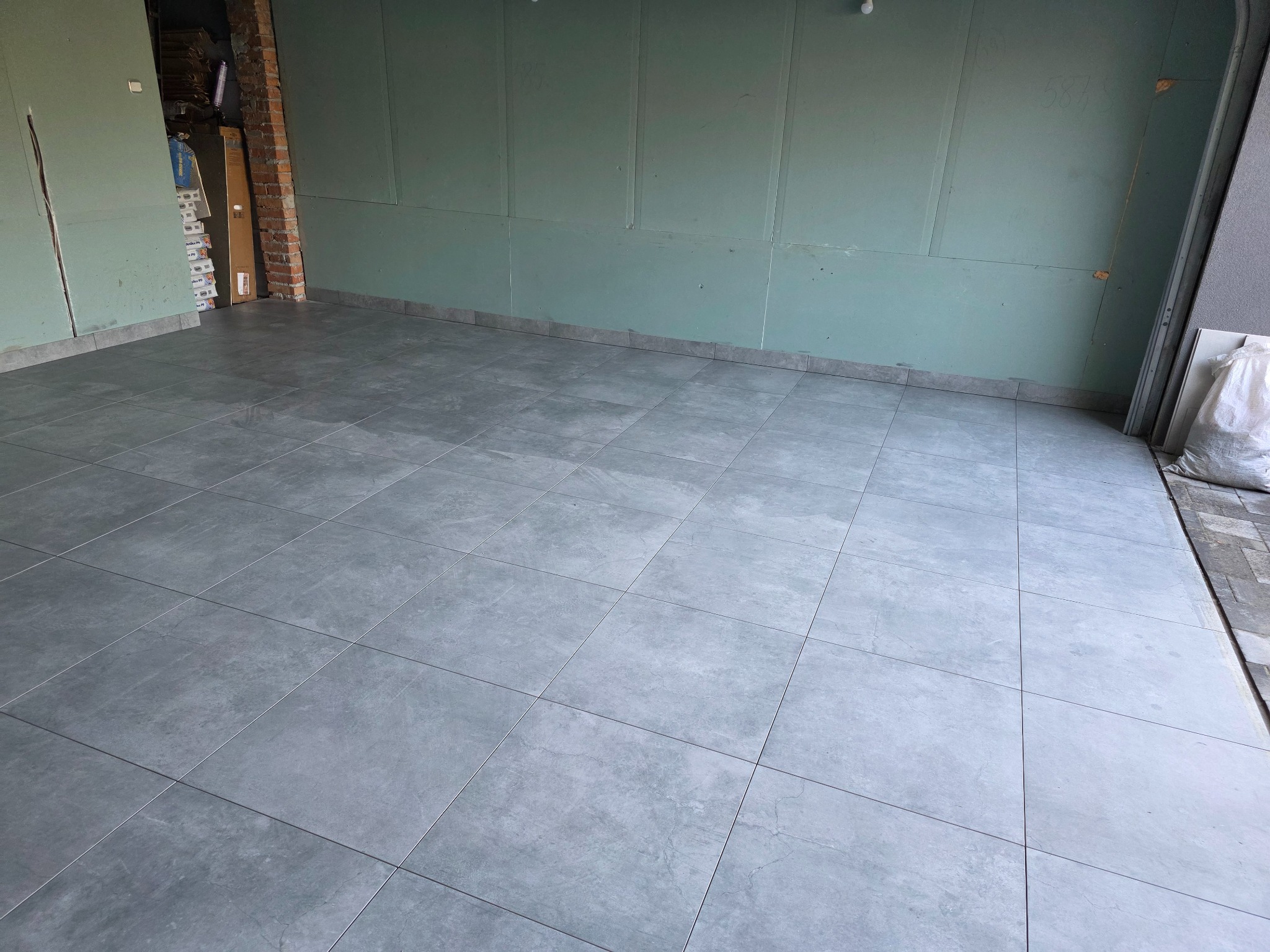 Szare płytki podłogowe ułożone w pomieszczeniu z ścianami wykończonymi płytami kartonowo-gipsowymi i szarą listwą przypodłogową. Częściowo widoczna cegła.