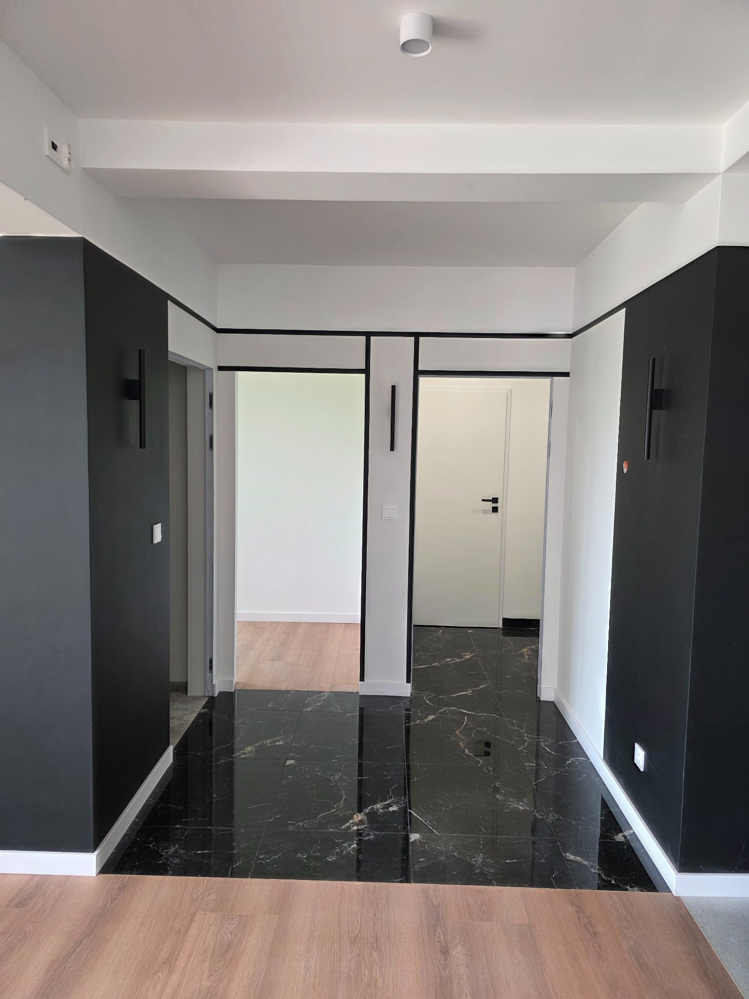 Hall z czarną marmurową podłogą, białymi ścianami z czarnymi akcentami i drewnianą podłogą w przejściach. Nowoczesne oświetlenie ścienne i sufitowe.