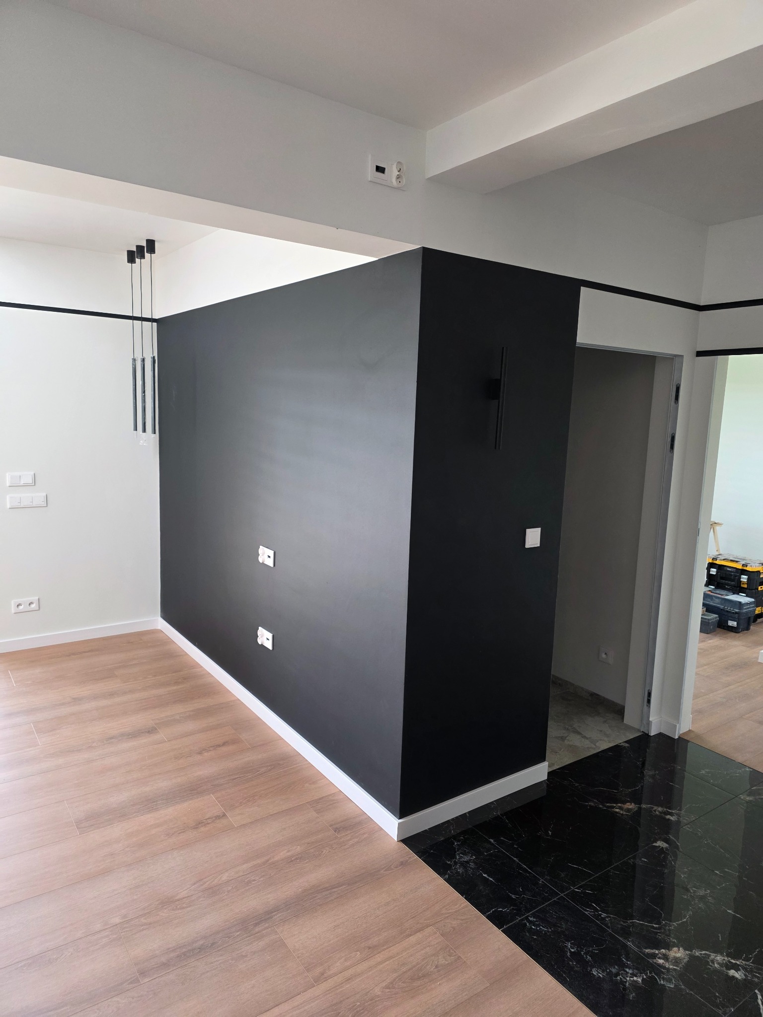 Nowoczesne wykończenie wnętrza: czarna ściana z gniazdkami, drewnopodobna podłoga, czarne płytki marmurowe i minimalistyczne oświetlenie.