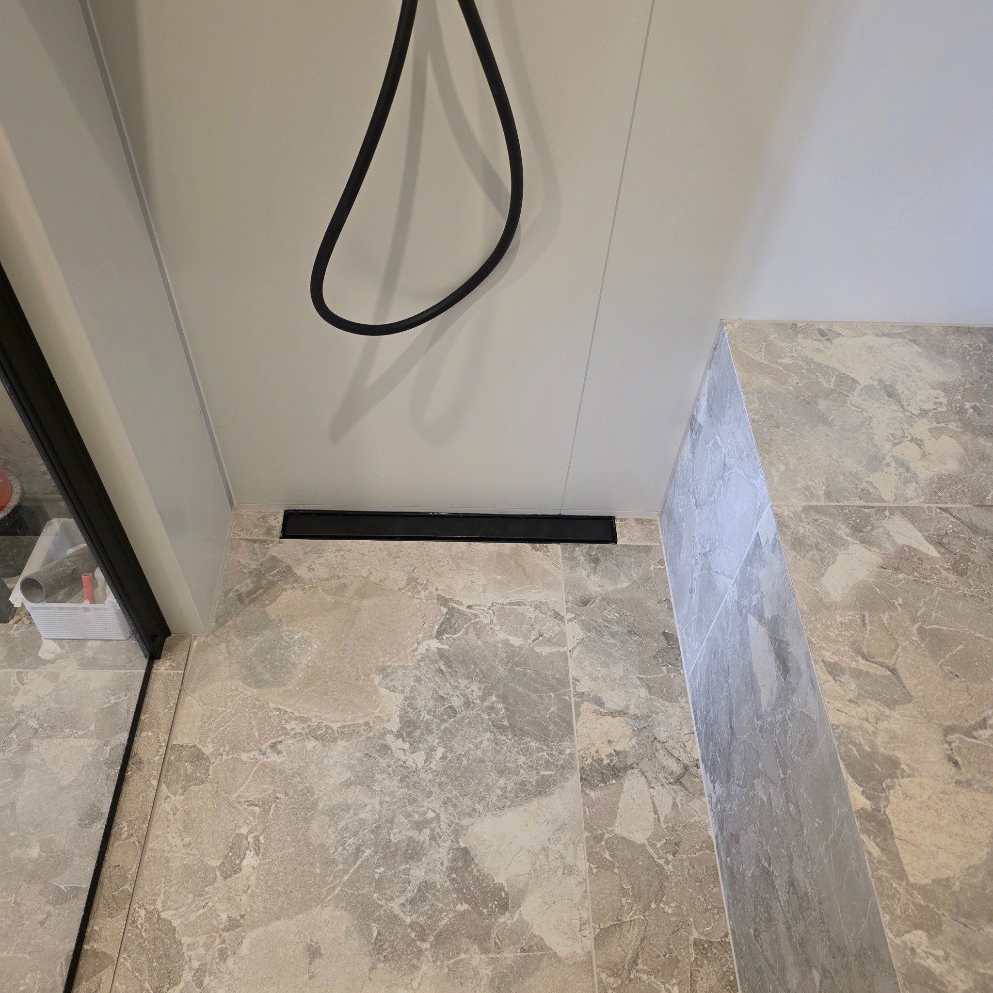 Wykończona łazienka z marmurową posadzką i odpływem liniowym. Widoczny fragment kabiny prysznicowej i czarny przewód prysznica na tle białej ściany.