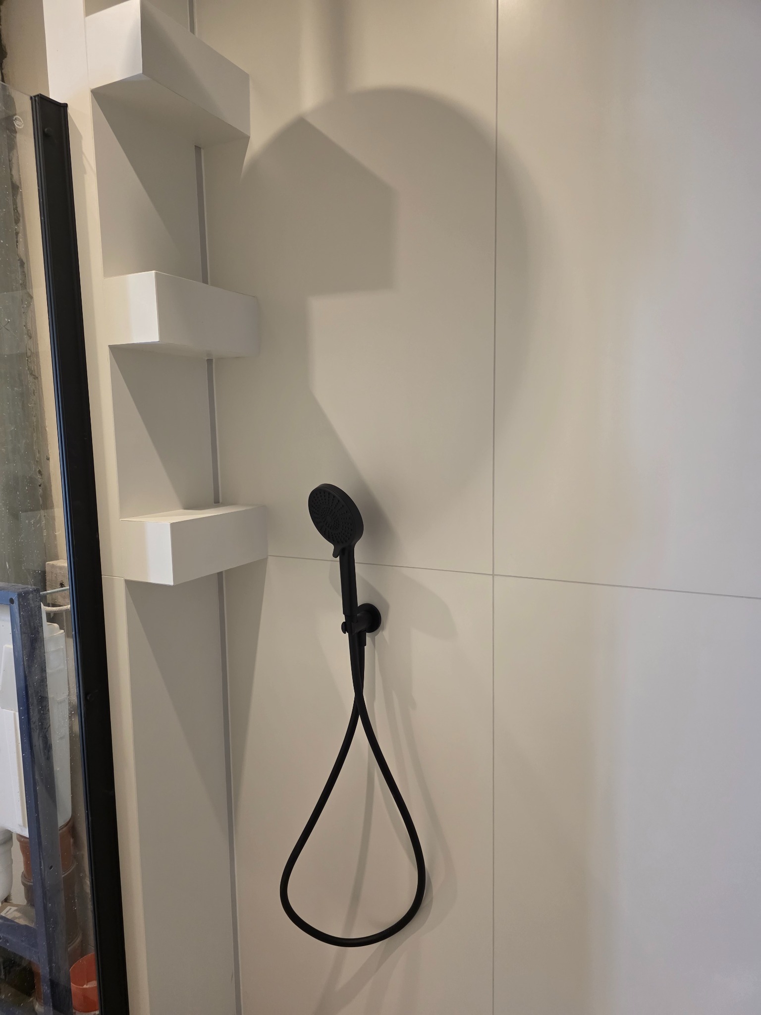 Minimalistyczna łazienka z białą armaturą: czarna słuchawka prysznicowa na tle białych płytek i półek. Nowoczesny design, widoczne elementy wykończeniowe.