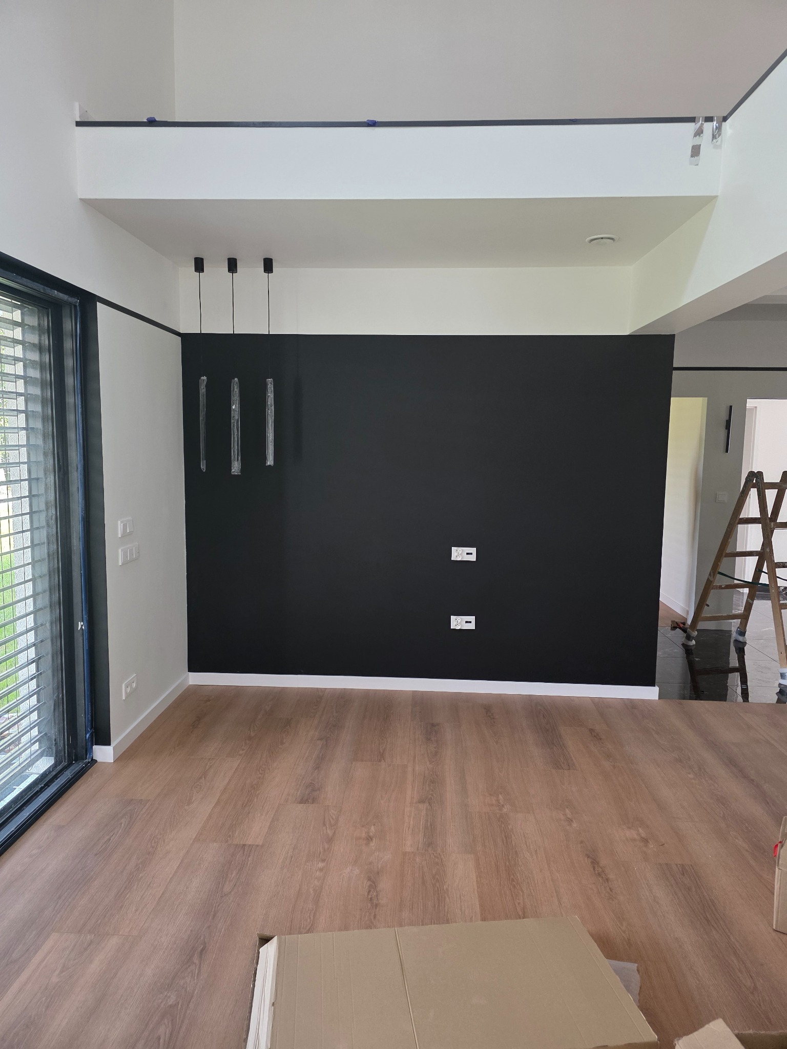Minimalistyczne wnętrze: czarna ściana kontrastuje z jasnymi ścianami i drewnianą podłogą. Widoczne nowoczesne lampy wiszące i drabina w tle.