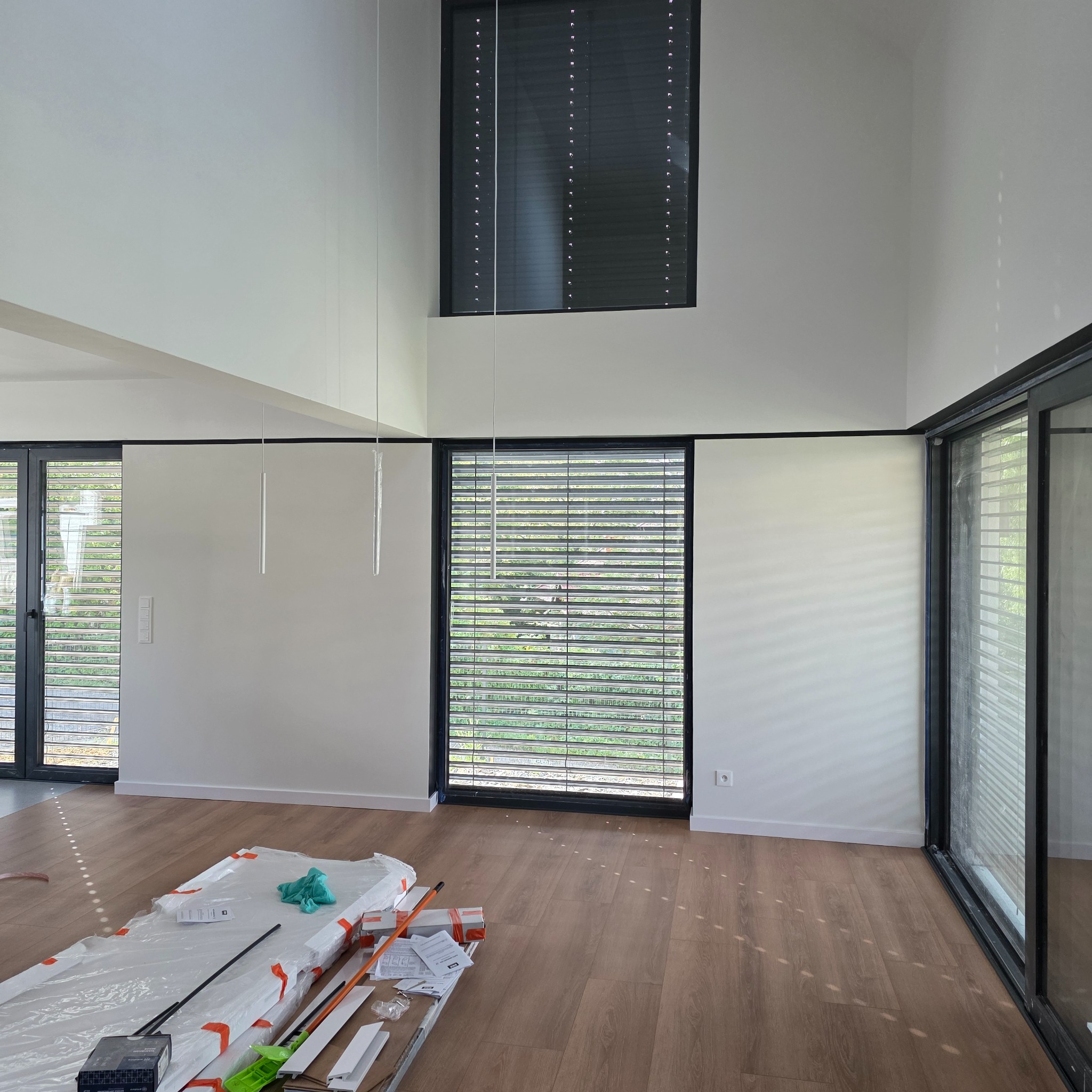 Jasne wnętrze z drewnianą podłogą i wysokimi oknami z żaluzjami. Na podłodze materiały budowlane, sugerujące prace wykończeniowe.