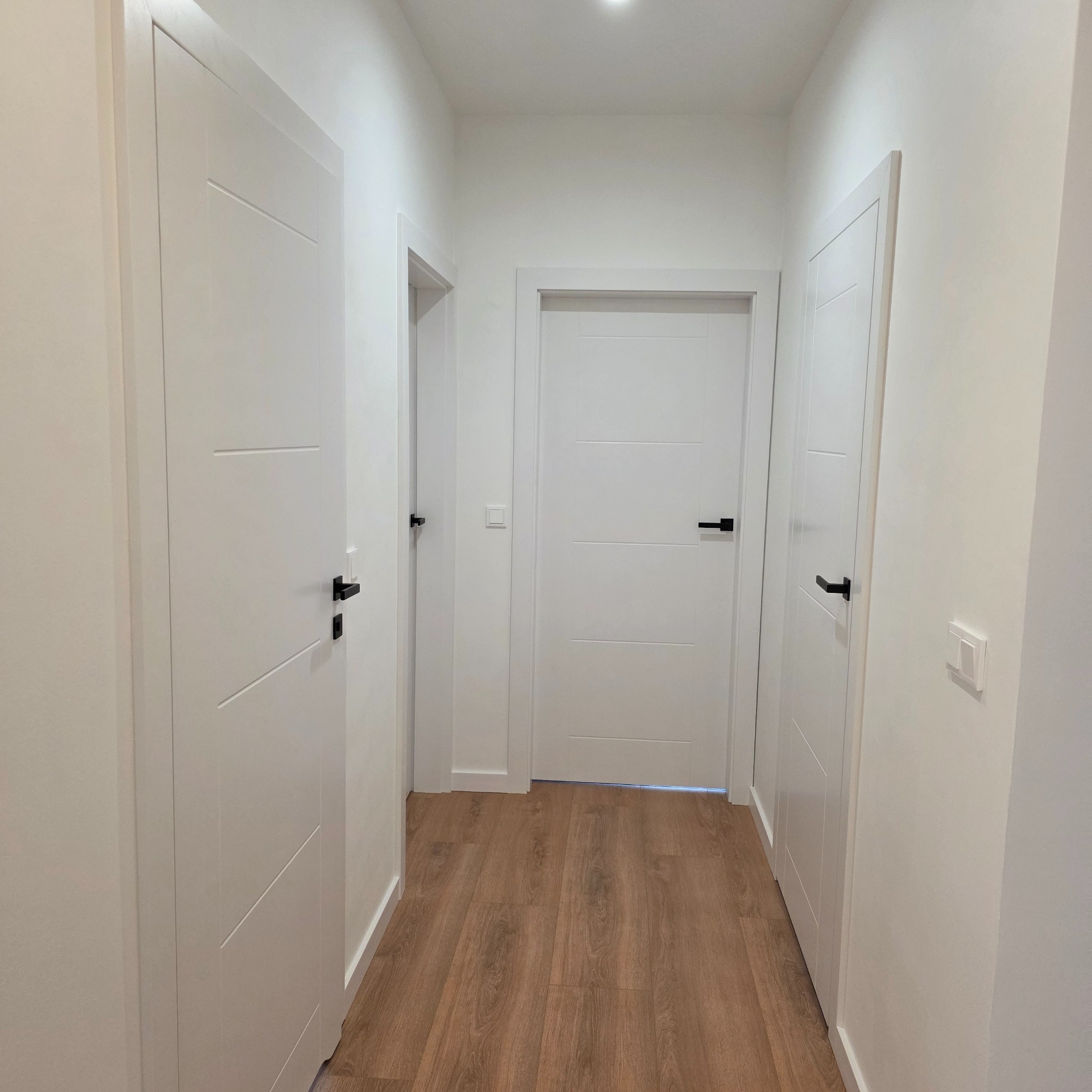 Jasne wnętrze korytarza po remoncie: białe ściany, drzwi z czarnymi klamkami i drewniana podłoga. Minimalistyczny design, świeże wykończenie.