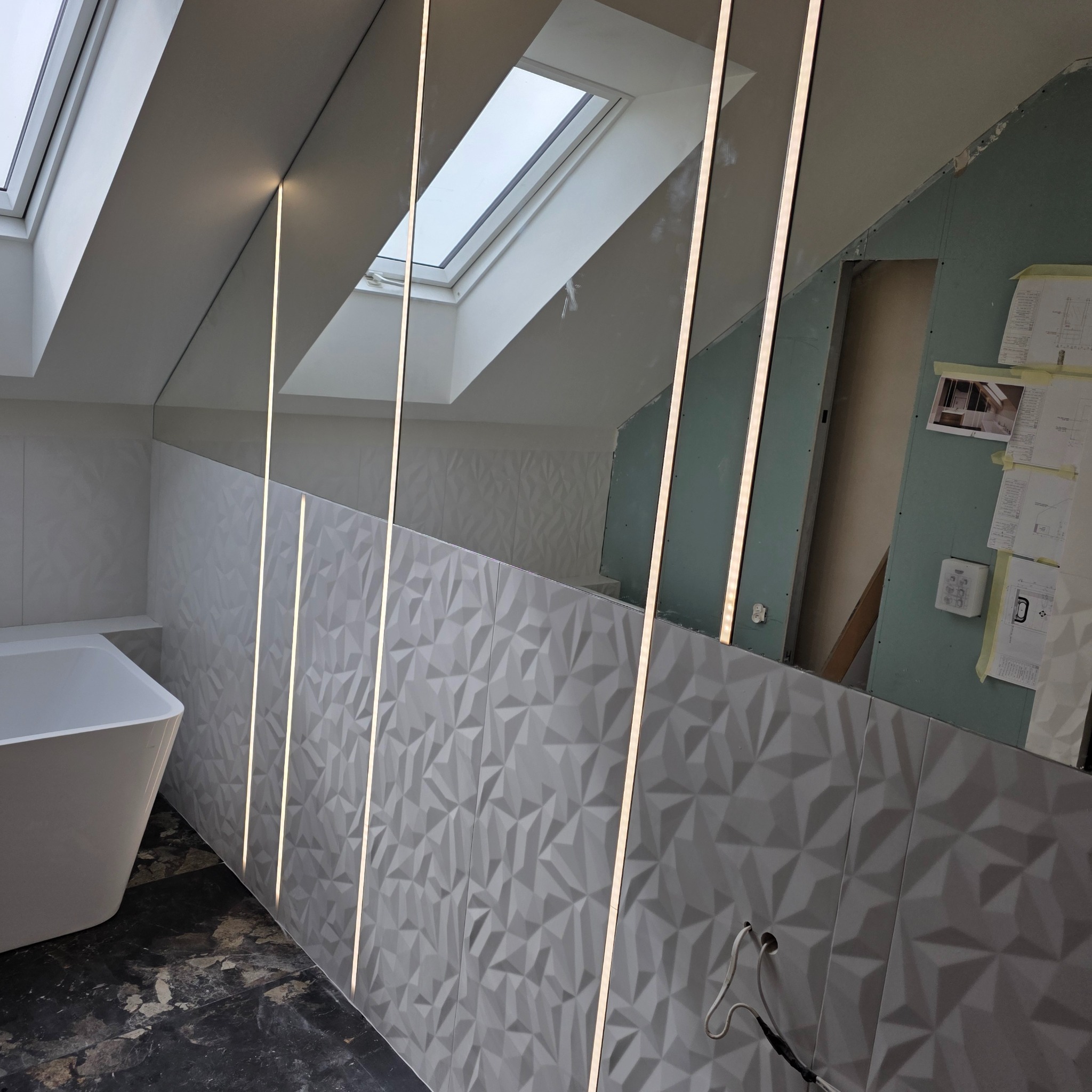 Nowoczesna łazienka na poddaszu z trójwymiarowymi płytkami, oświetleniem LED i oknem dachowym. Widoczne fragmenty wanny i ściany w trakcie wykończenia.