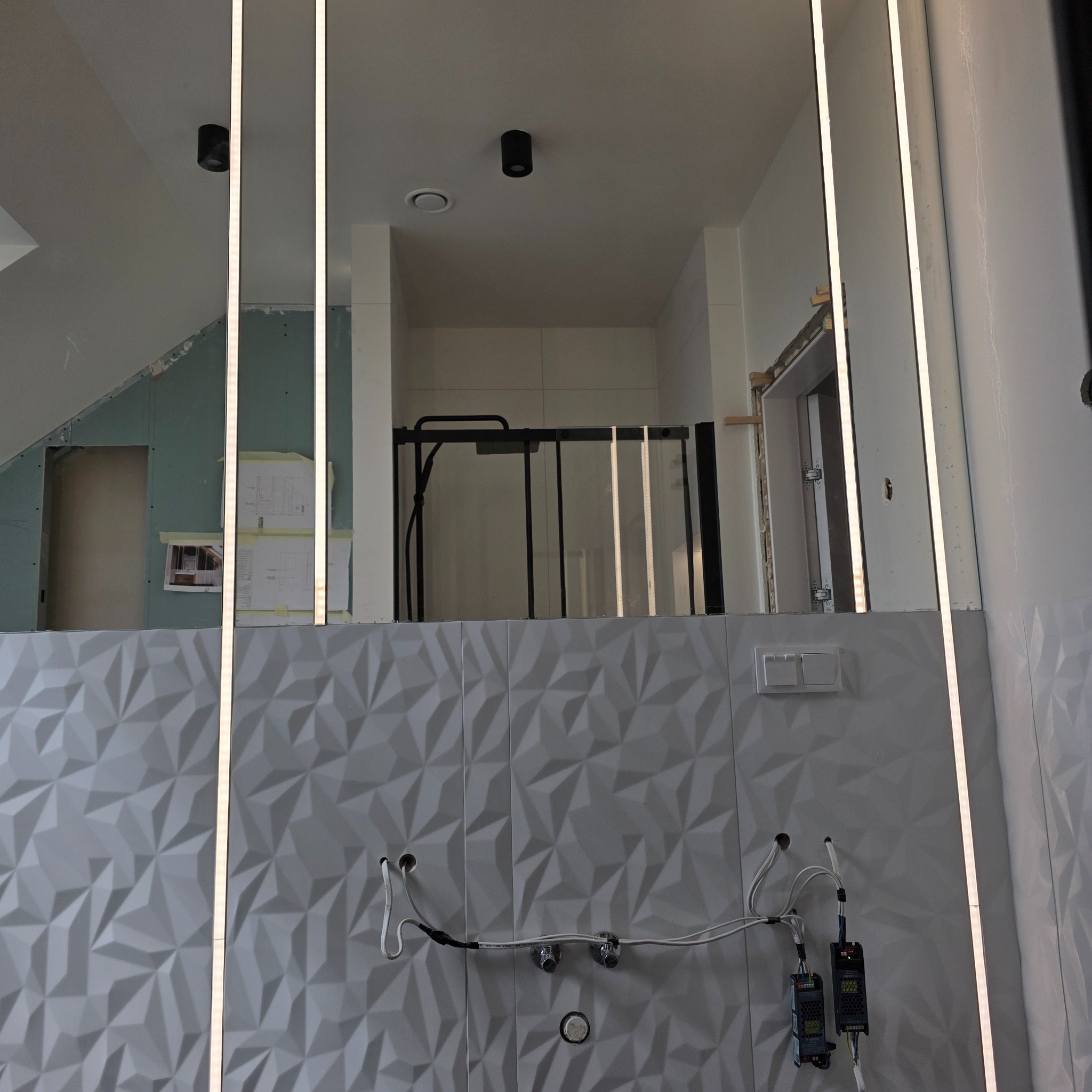 Łazienka w trakcie wykańczania: białe, trójwymiarowe płytki, lustro z oświetleniem LED, widoczne instalacje i fragmenty kabiny prysznicowej. W tle płyta karton-gips.