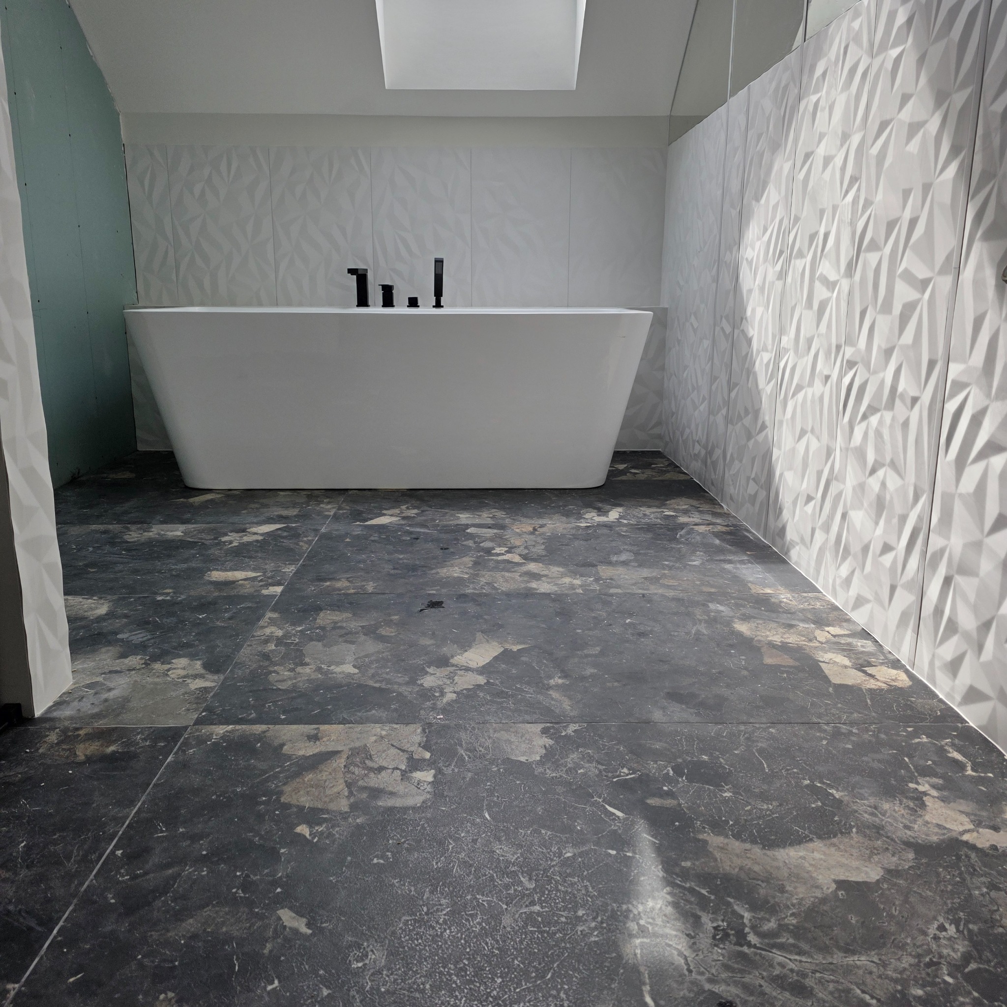 Nowoczesna łazienka z wolnostojącą wanną na tle trójwymiarowych, białych płytek ściennych i ciemnej, marmurkowej podłogi. Widoczna surowa ściana z płyty gipsowej.