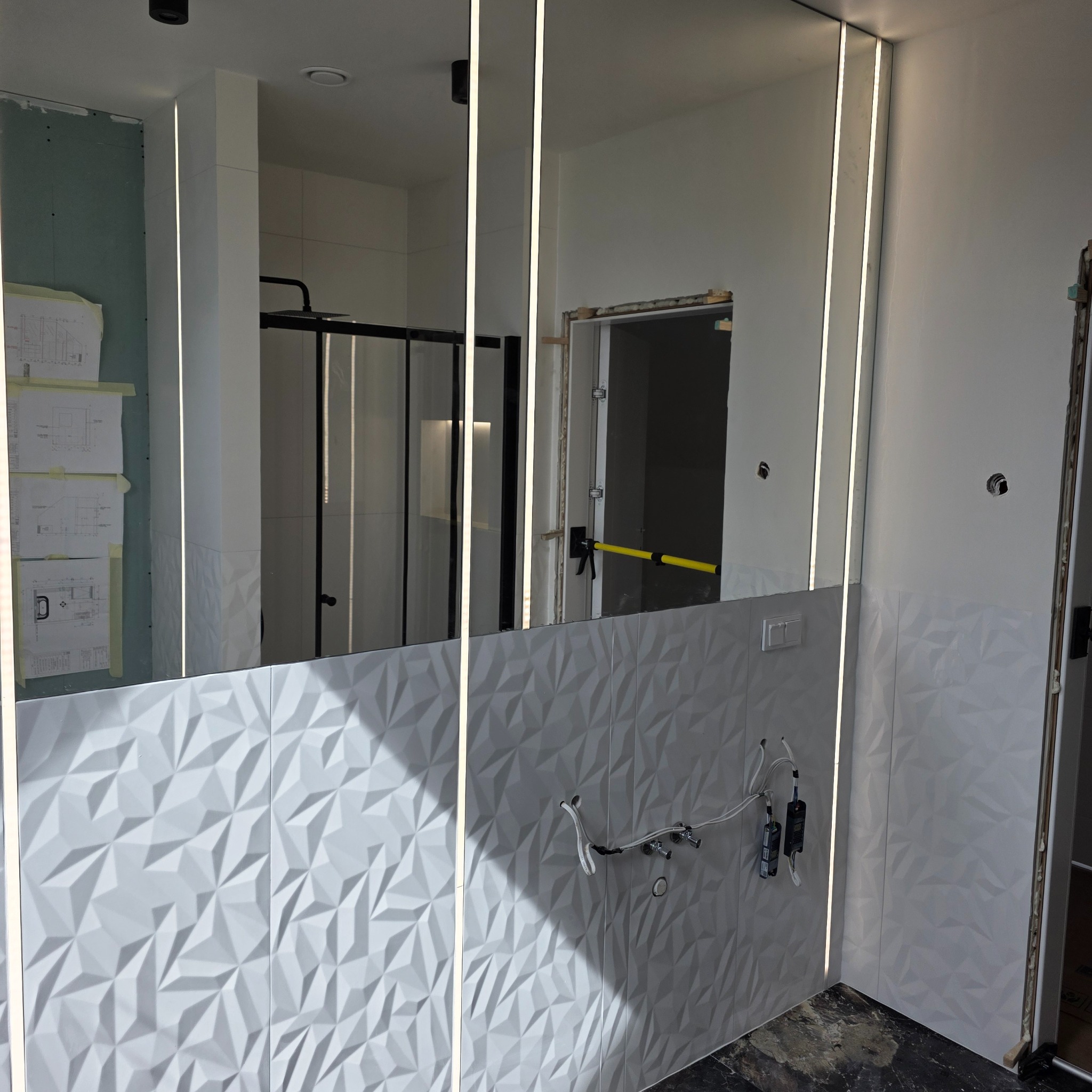 Nowoczesna łazienka w trakcie wykańczania z podświetlanym lustrem i trójwymiarowymi płytkami. Widoczne niedokończone instalacje.