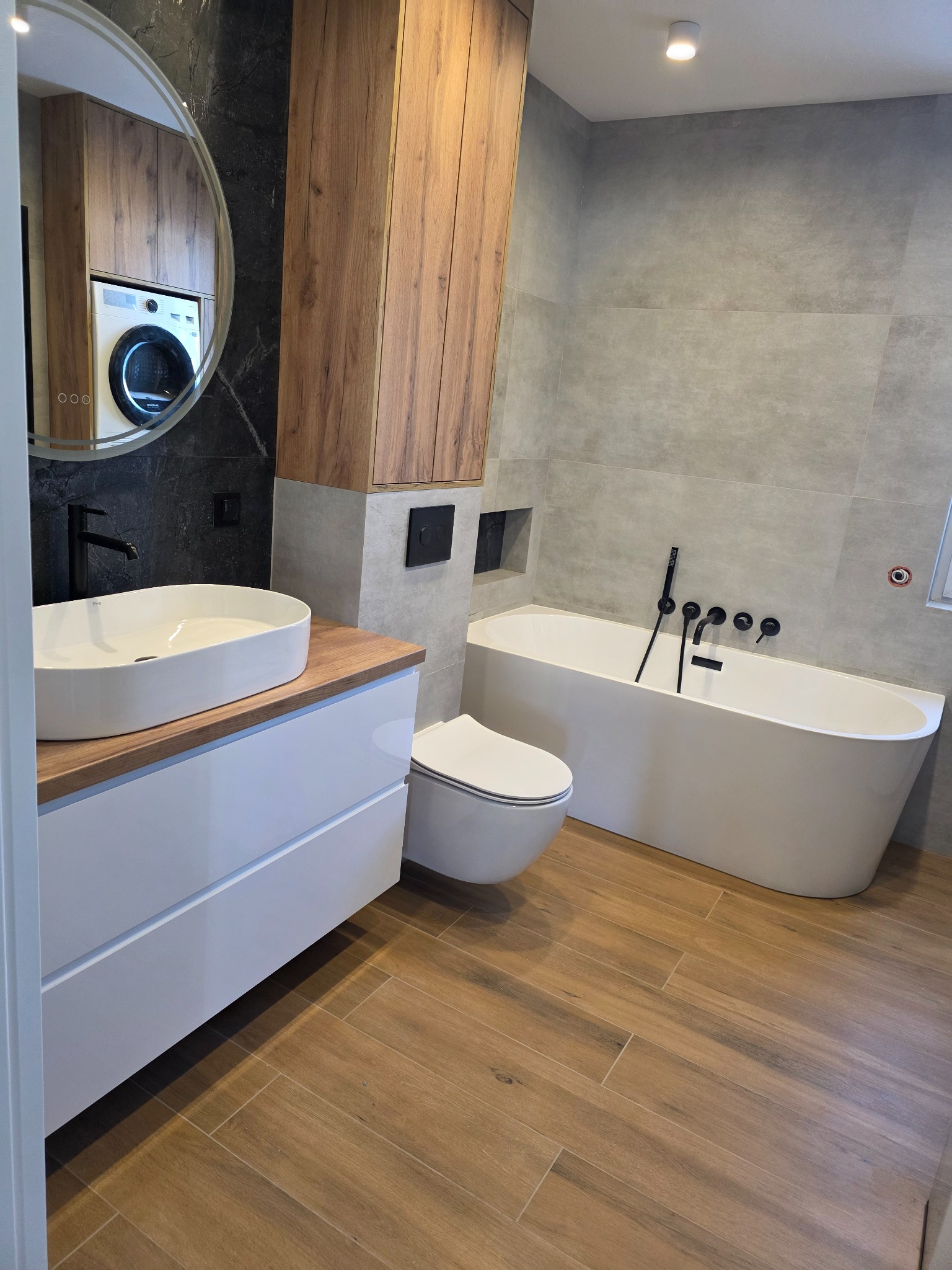 Nowoczesna łazienka w Krakowie z wanną, umywalką na szafce i drewnopodobną podłogą. W lustrze odbija się pralka. Minimalistyczny design z czarnymi akcentami.