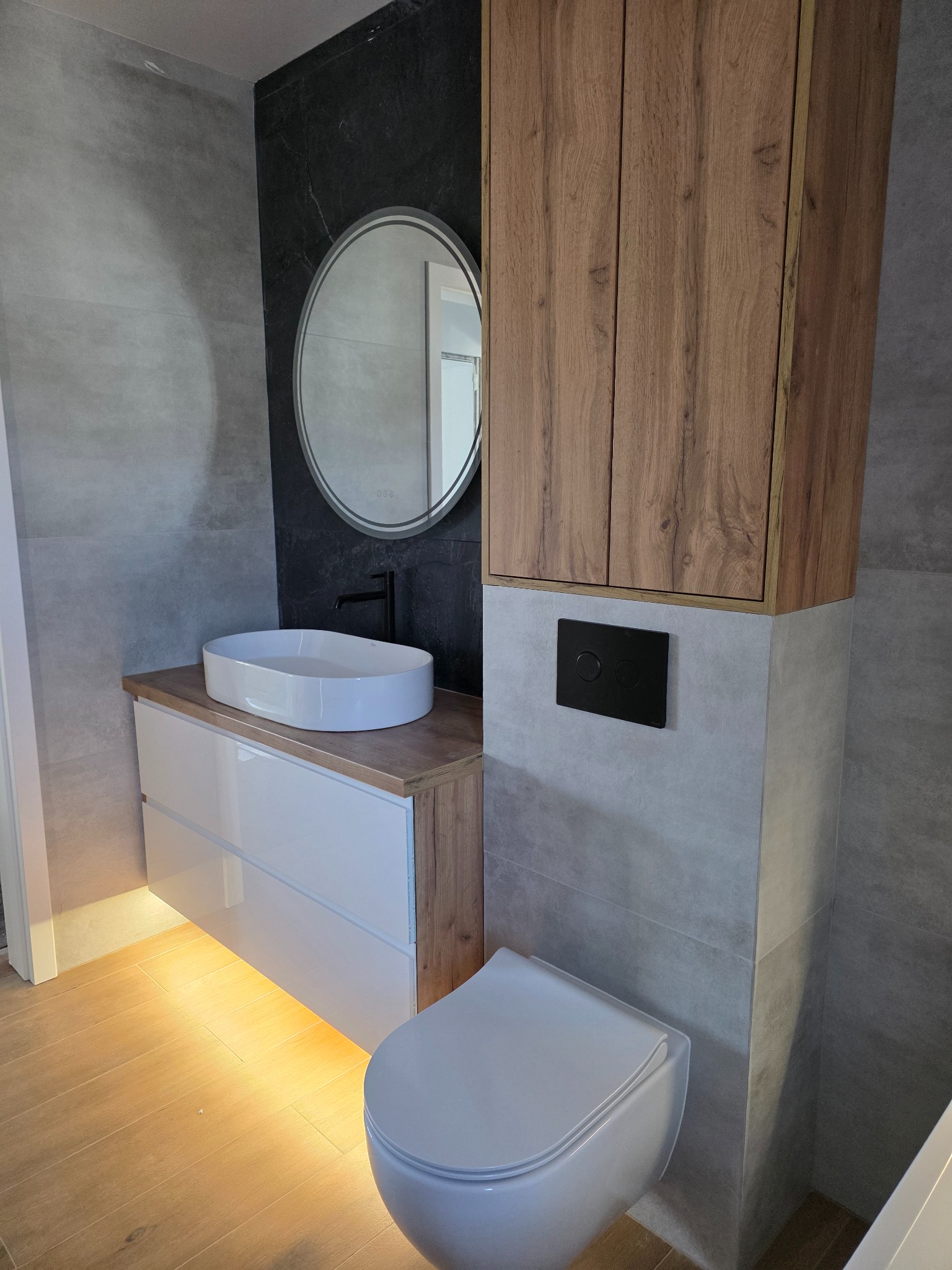 Nowoczesna łazienka po remoncie: biała umywalka nablatowa, okrągłe lustro z podświetleniem, drewniane szafki i szara płytka. Podświetlenie LED pod szafką dodaje ciepła.