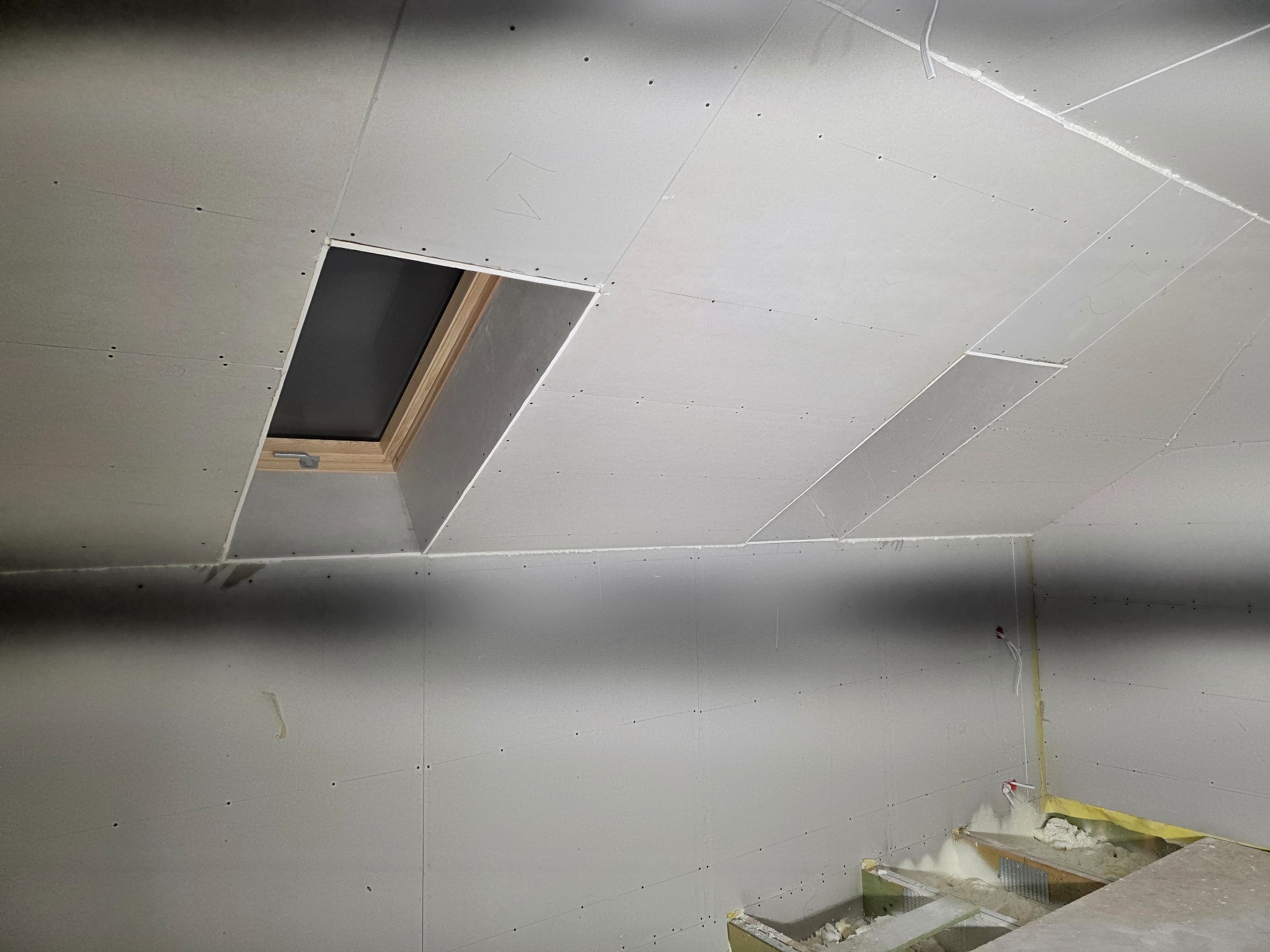Montaż płyt gipsowo-kartonowych na poddaszu z oknem dachowym. Widoczne łączenia płyt i wkręty. Częściowo ocieplone poddasze. Ujęcie z dołu.