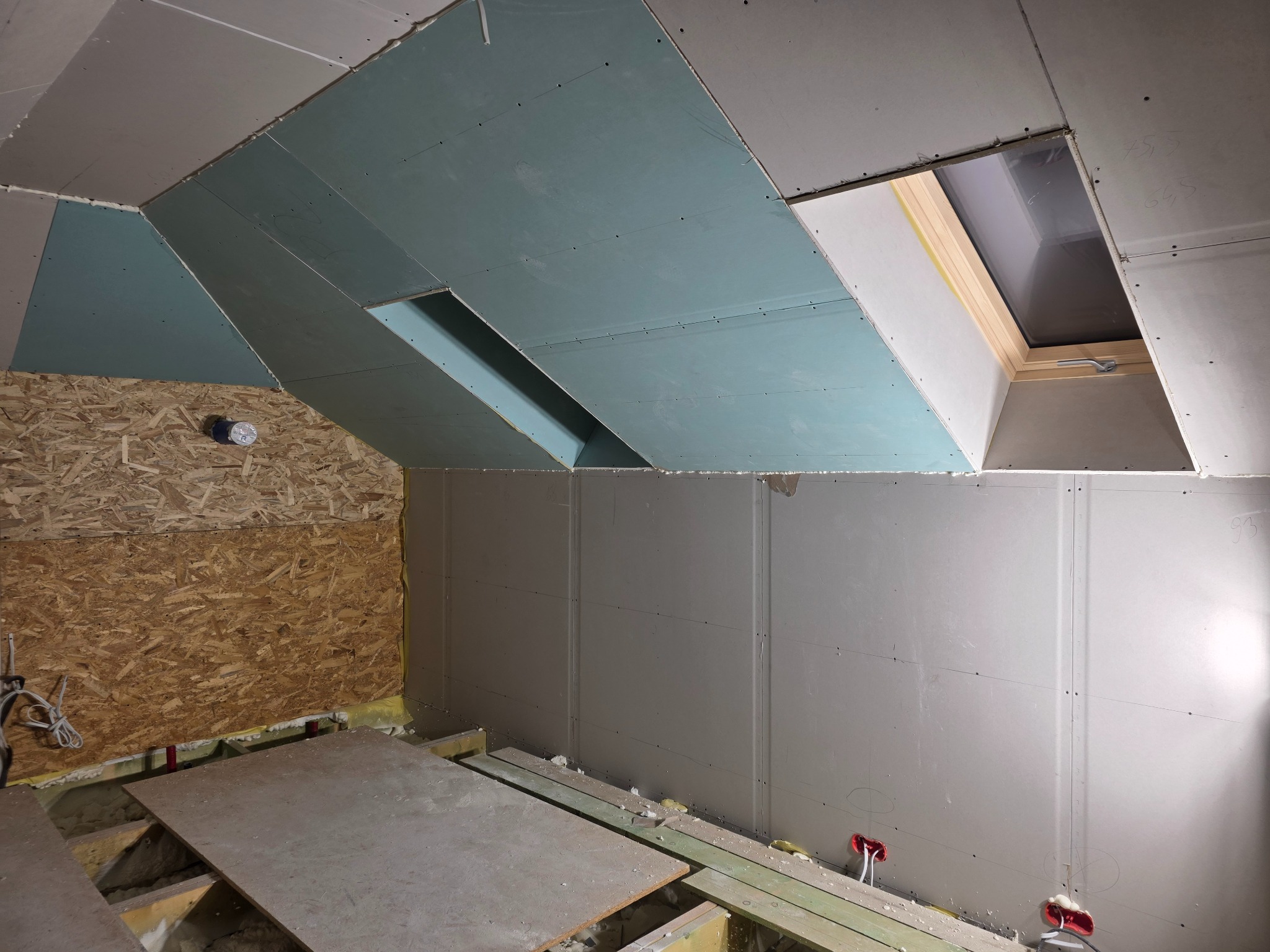 Montaż płyt karton-gips na poddaszu ze skośnym dachem i oknem dachowym. Widoczne płyty GK w kolorze szarym i zielonym, drewniana ściana z płyty OSB i fragment podłogi.