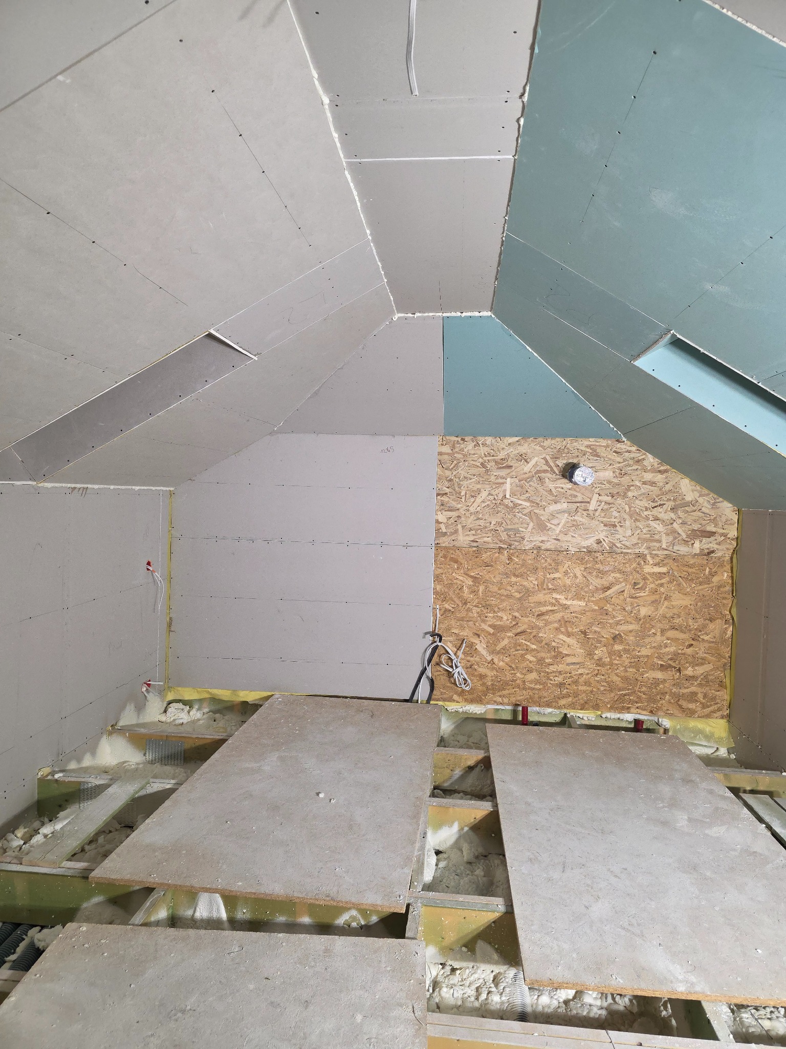 Skośny strych w trakcie zabudowy płytami gipsowo-kartonowymi. Widoczne ocieplenie, fragmenty instalacji elektrycznej i płyty OSB. Prace wykończeniowe w surowym stanie.